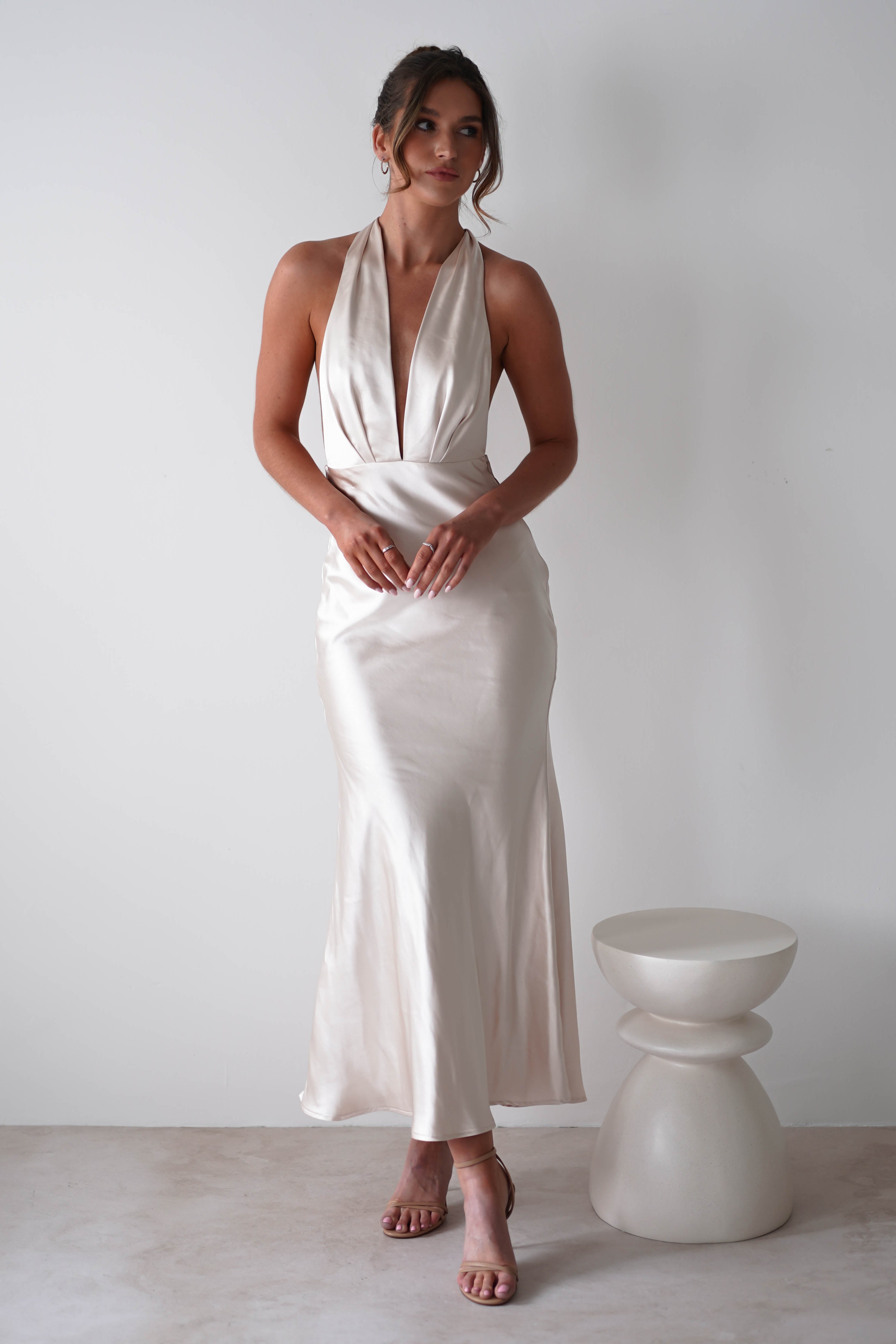 A woman stands against a white wall wearing a sleeveless, ivory gown crafted from soft satin material with a deep V-neck and open back. Her hair is up, strappy heels complete the look, and a modern sculptural stool sits beside her—perfect for rehearsal dinners.