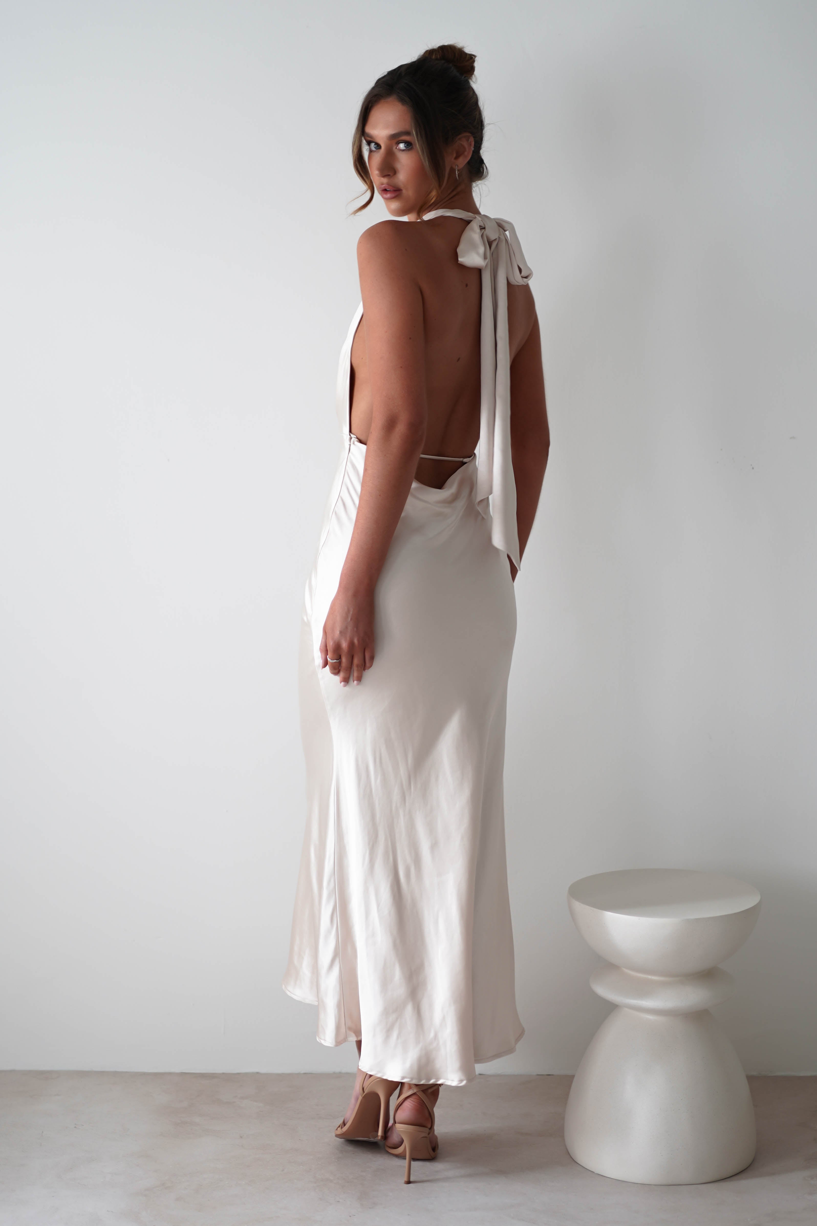 Woman in a backless, floor-length white satin dress with a glamour neck-tie bow stands barefoot against a plain white wall, next to a small white sculptural stool—perfect elegance for rehearsal dinners.