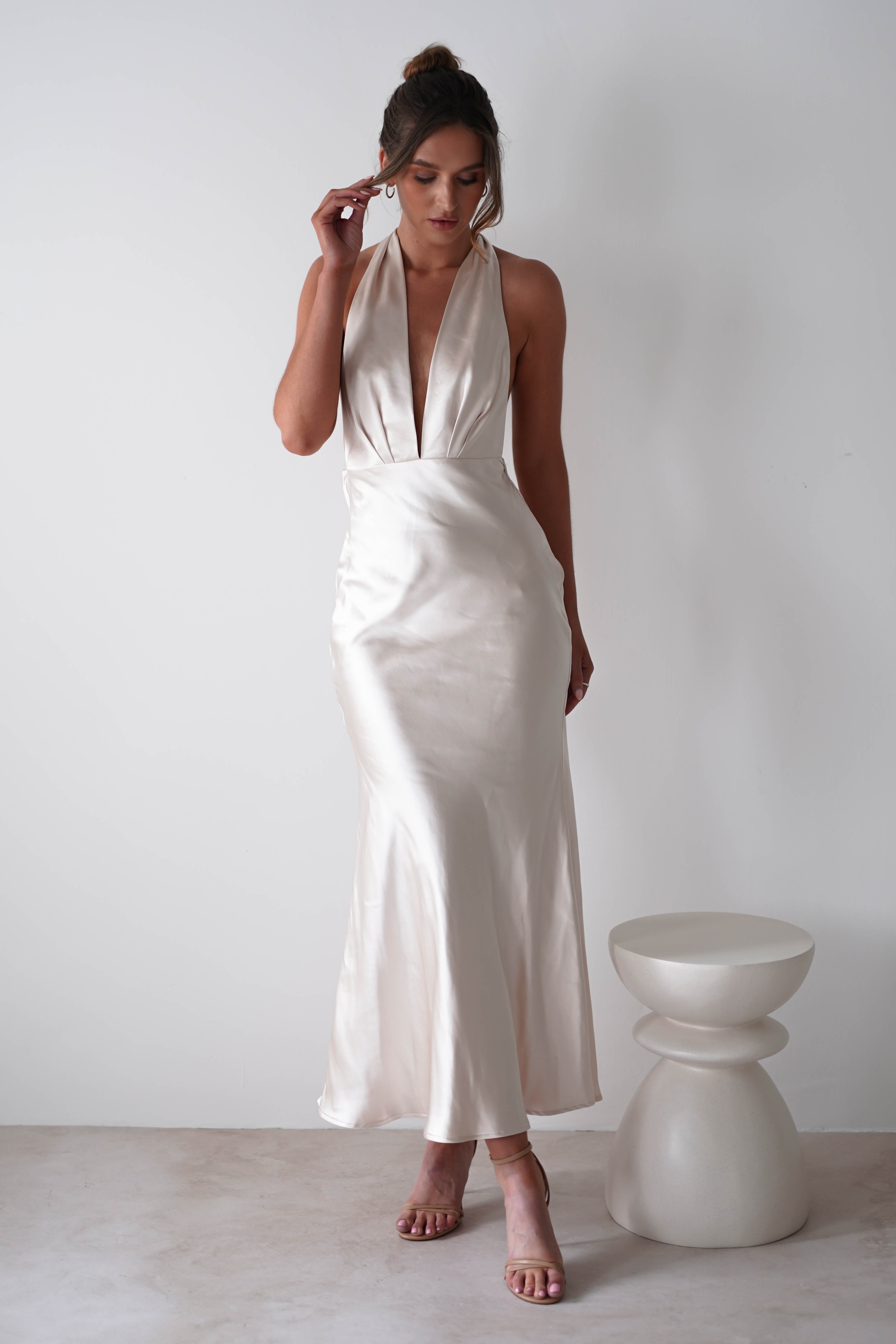 A woman in a silky, ivory halter-neck gown crafted from soft satin material stands beside a modern white stool, posing against a plain white wall. She has her hair in a loose bun and wears strappy nude heels—perfect for rehearsal dinners.