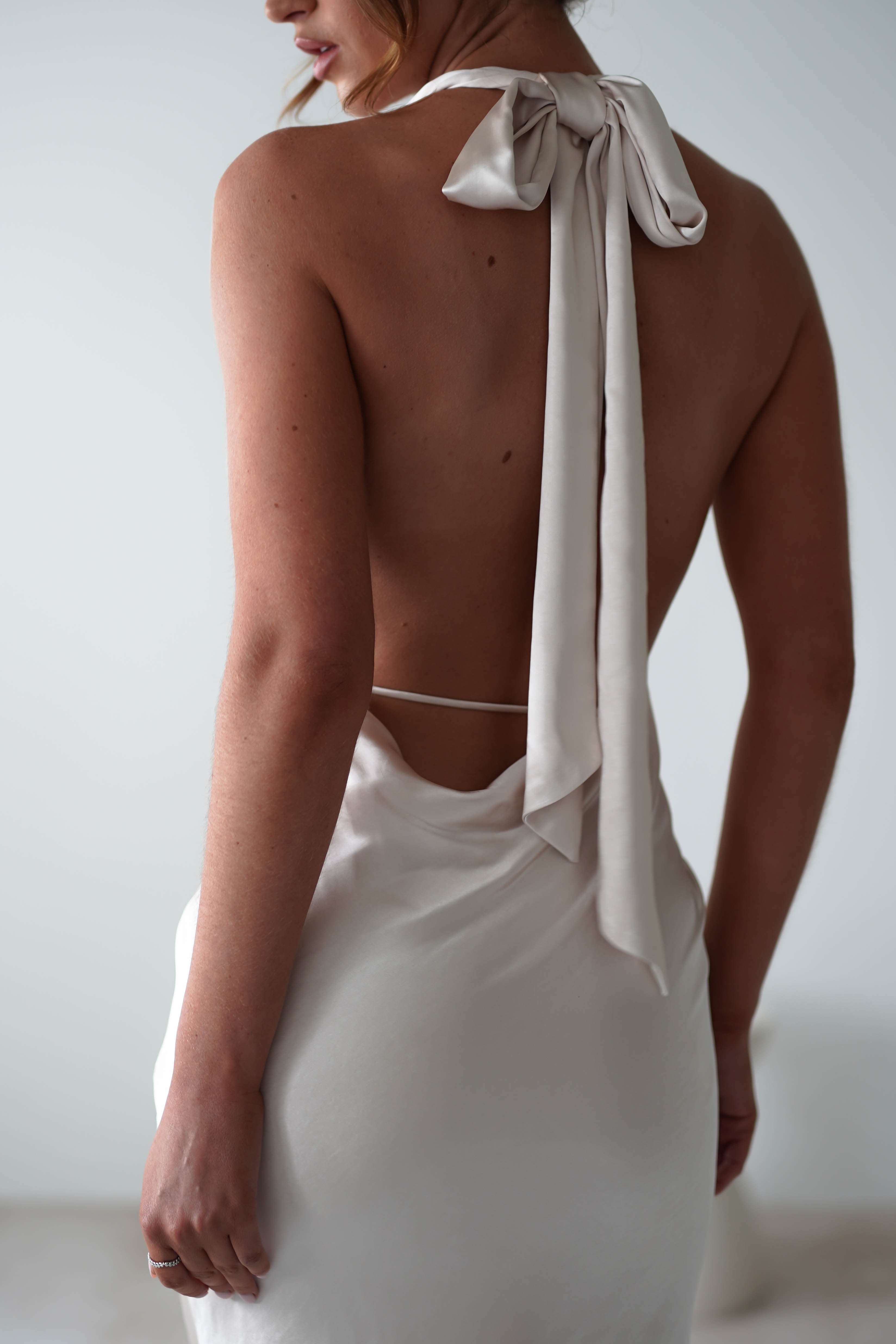 A woman wearing a backless, light-colored dress crafted from soft satin material with a glamour neck-tie bow at the neck, shown from behind against a plain background—perfect for rehearsal dinners.