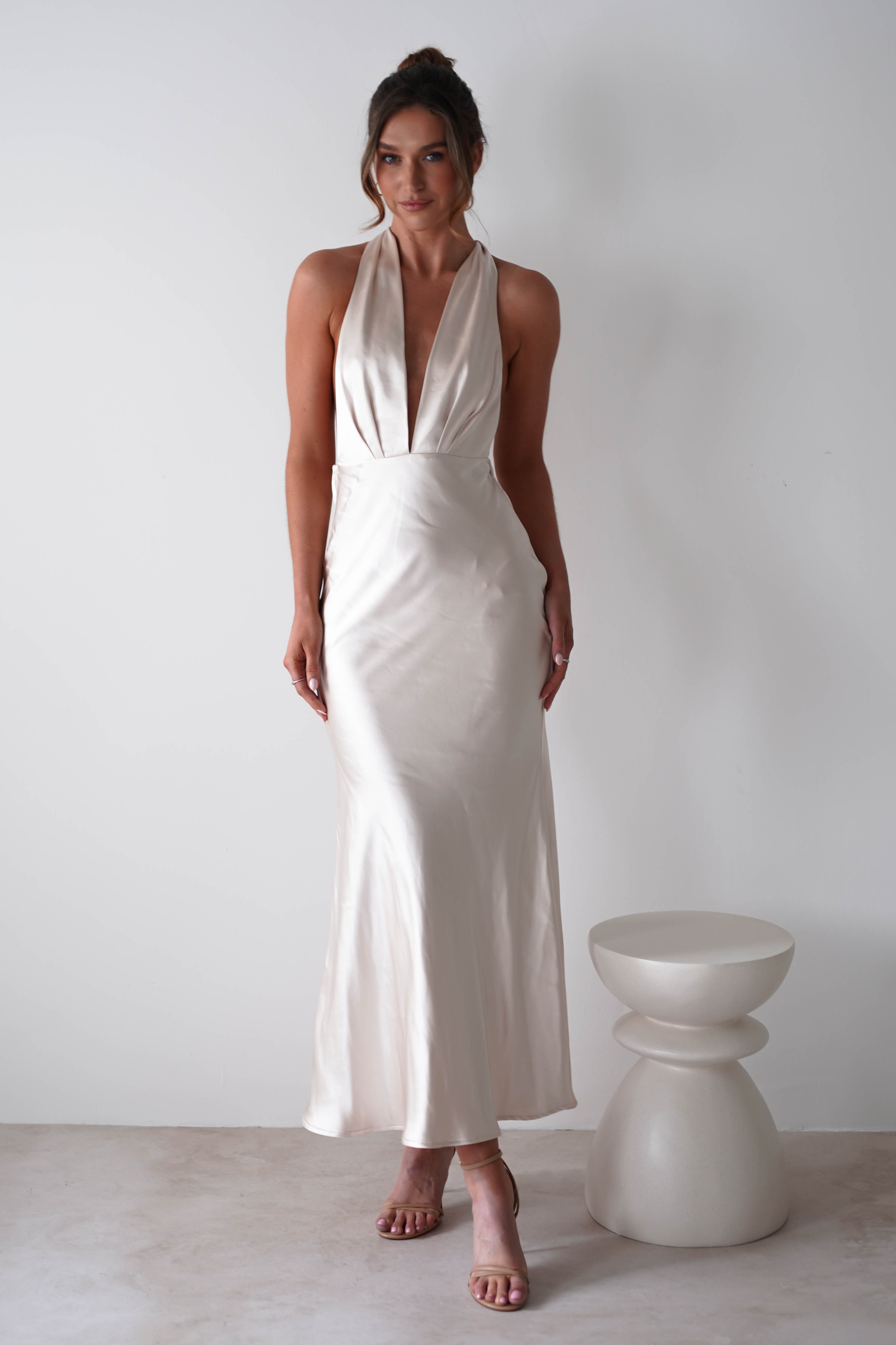 Woman wearing a white, sleeveless satin halter dress with a glamour neck-tie and deep V-neckline, standing next to a modern white stool against a plain white background. Perfect for rehearsal dinners or elegant events.