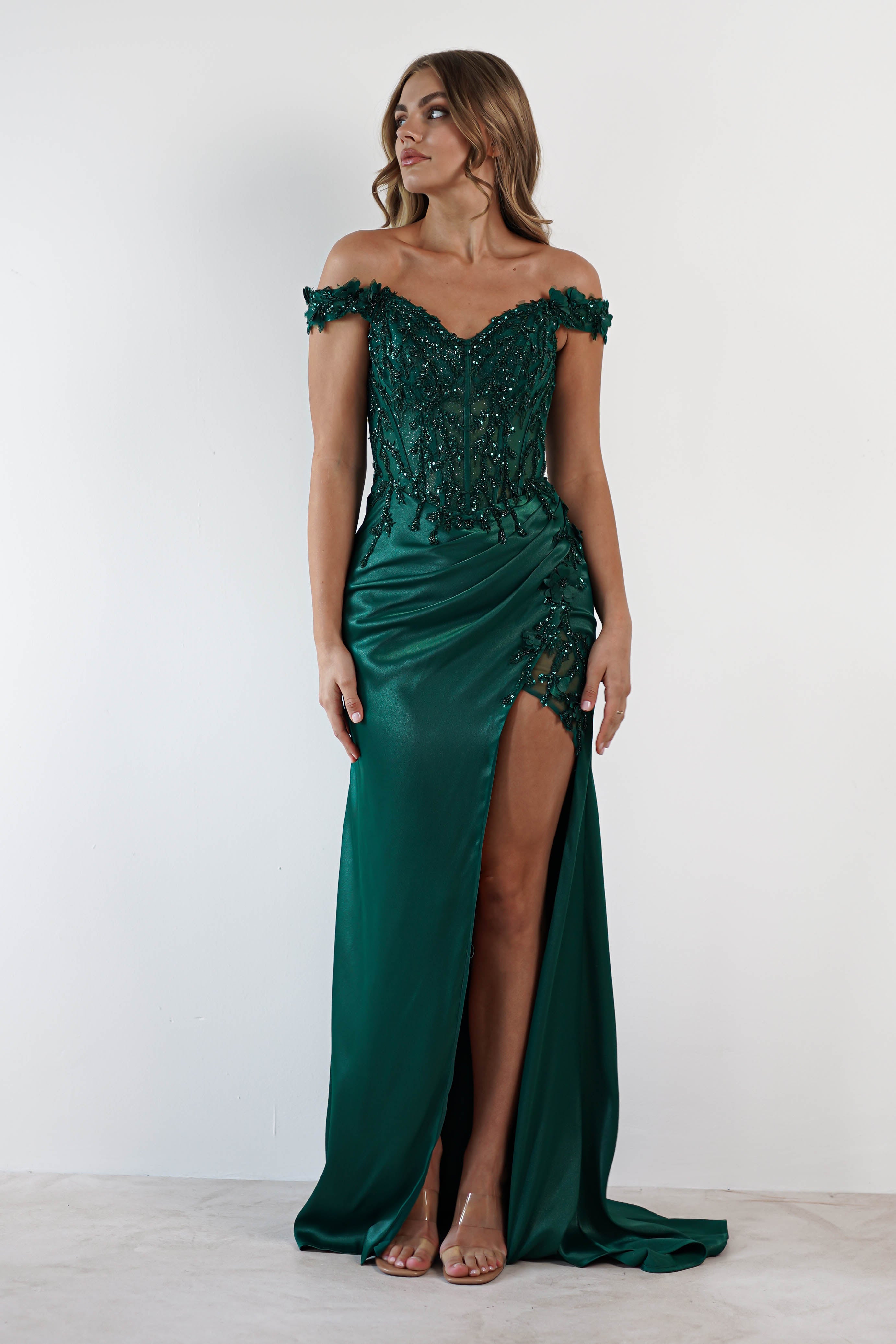 A woman in an off-shoulder, emerald green satin gown with intricate beading stands against a white background. The dress features a high slit and fitted bodice with a lace-up back. She gazes to the side, wavy hair cascading over her shoulders, and wears clear heels.
