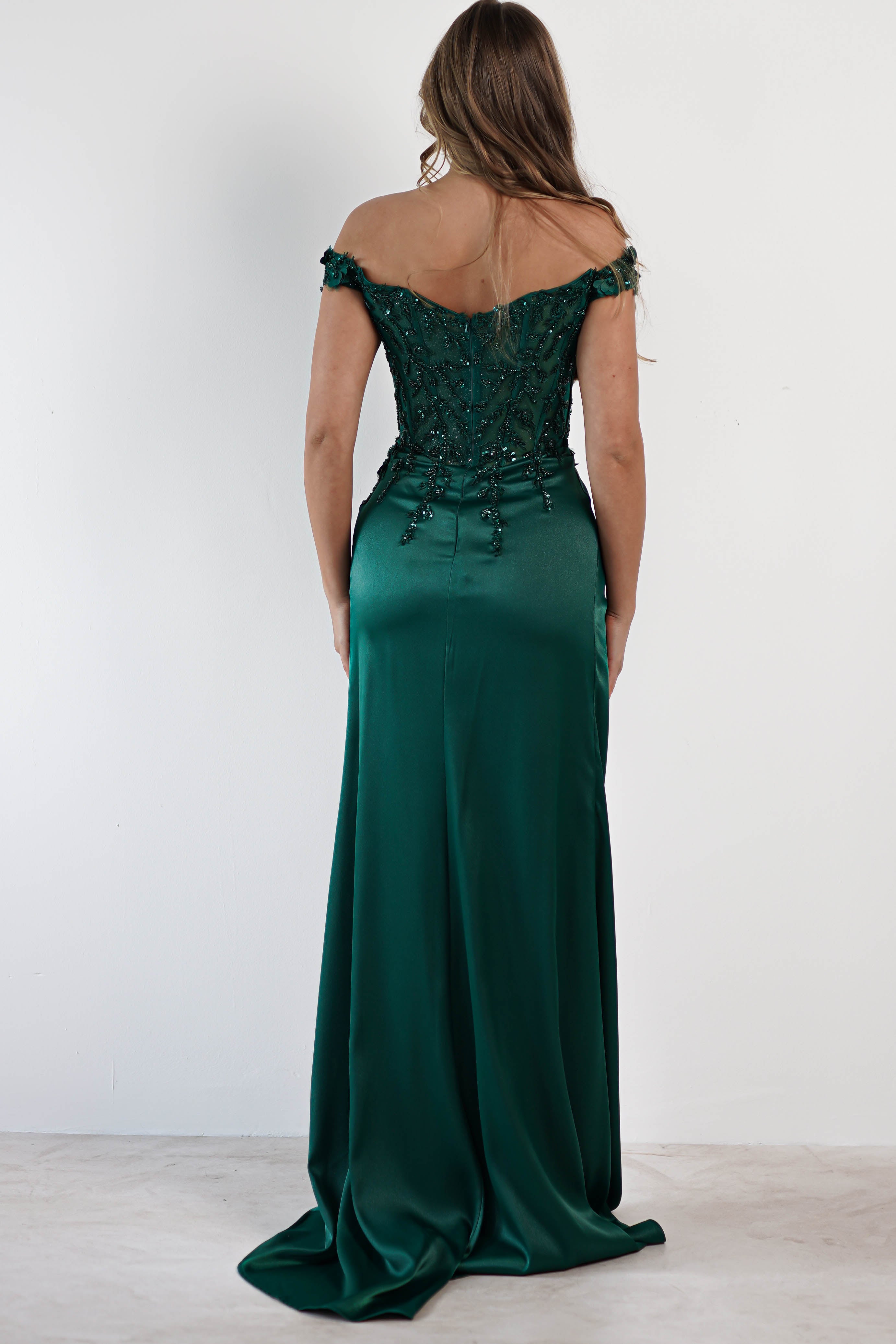 A woman stands facing away, wearing a stunning satin gown in emerald green, boasting intricate beading on the bodice. Her brown hair cascades down her back as the gown flows elegantly to the floor with a daring high slit, adding a touch of allure to her ensemble.