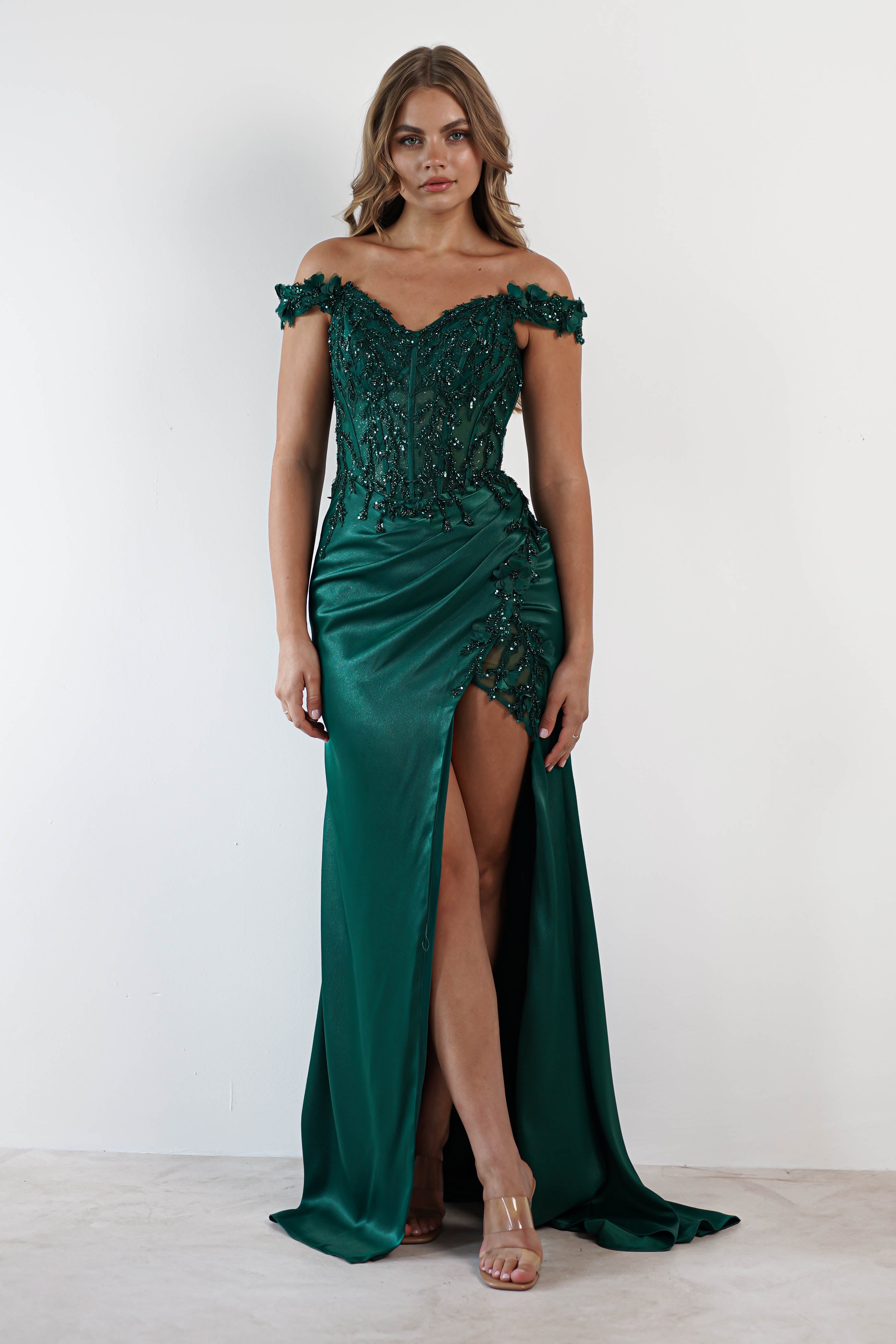 A woman in a satin emerald green gown featuring a sweetheart neckline and intricate beading stands against a plain white background, her attire elegantly completed with a daring thigh-high slit.