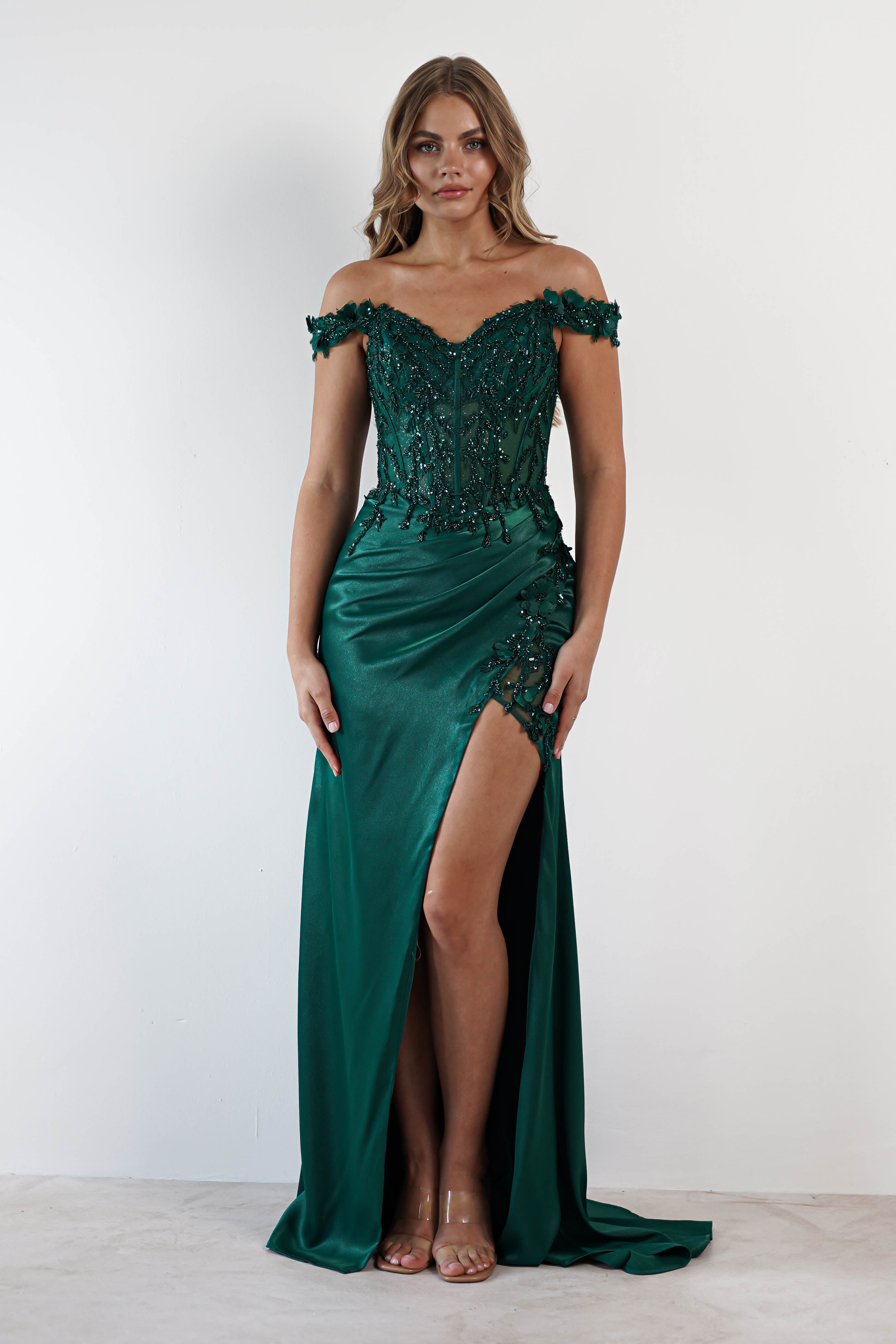 A woman stands wearing an elegant, off-the-shoulder emerald green gown with intricate beadwork on the bodice and a sultry high slit. The satin gown flows gracefully as she poses against a plain white background, looking directly at the camera.