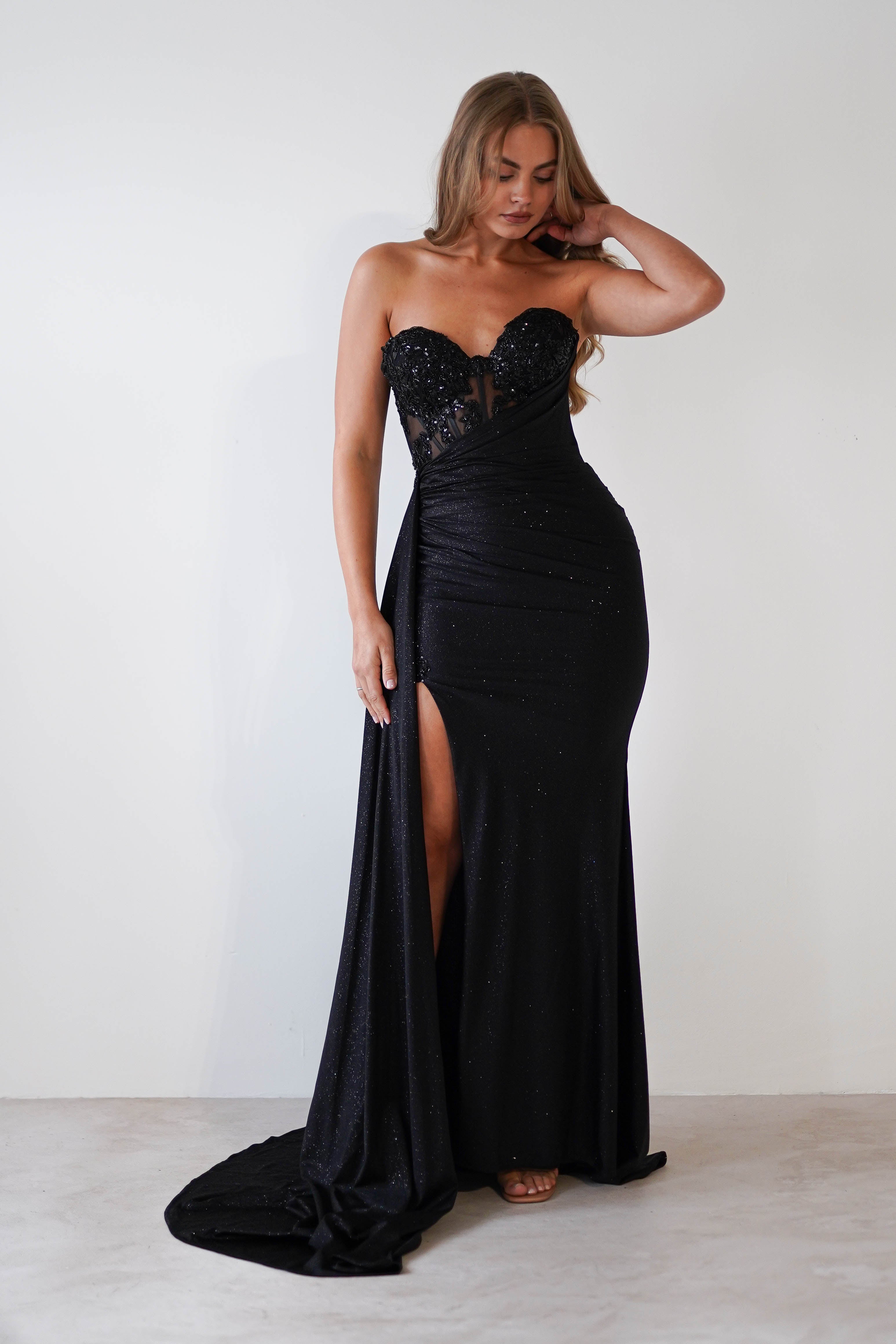 A person poses in a strapless black gown with a sweetheart neckline and a high slit. The glitter gown is embellished with sparkling details, and they have long wavy hair. They stand against a plain white background, perfect attire for formal events.