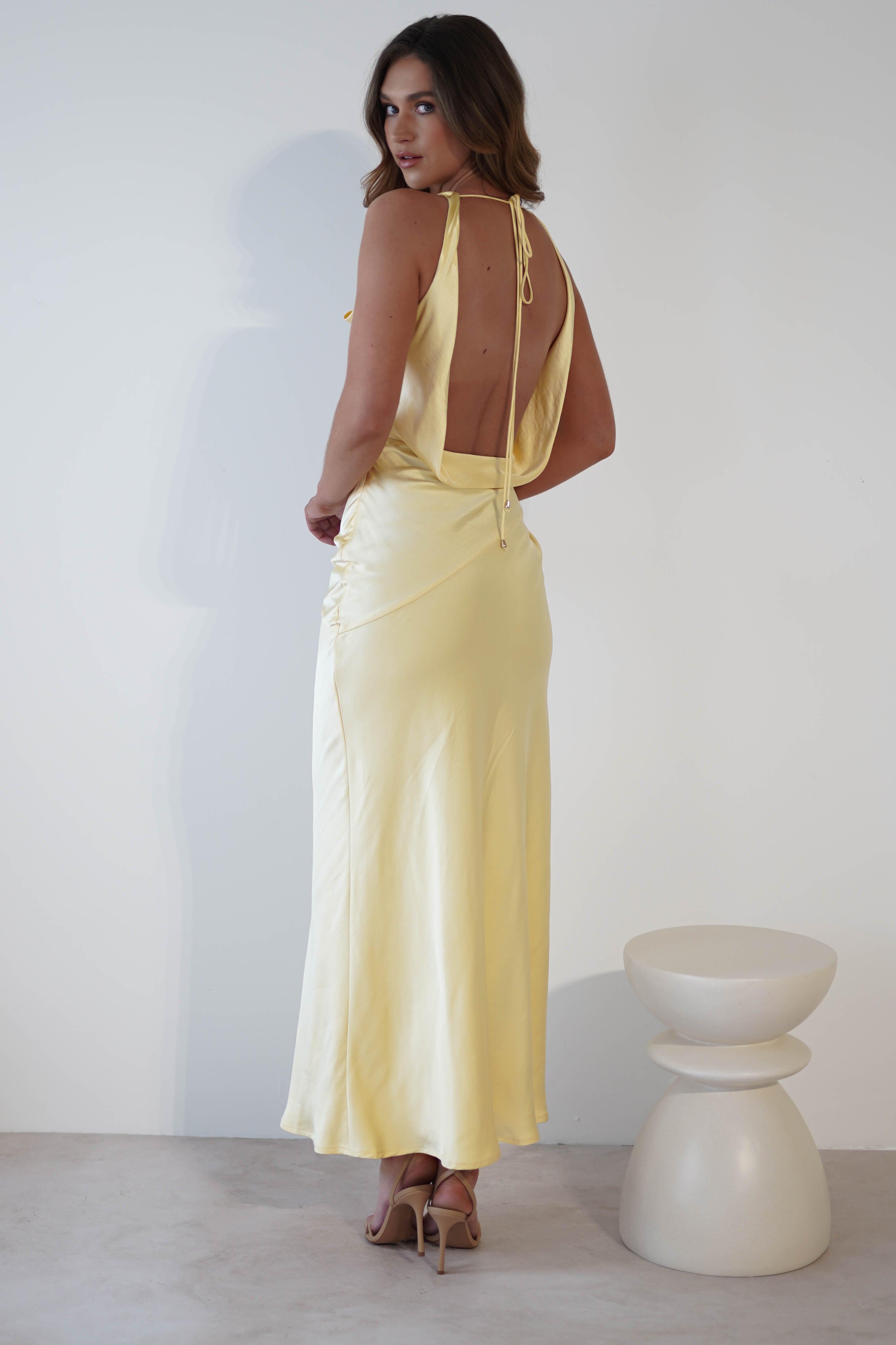 A woman in a light yellow satin gown with a cowl neckline and draped open-back stands on a light-colored floor. Facing sideways with elegance, she pairs high heels with her look. A plain white wall and small round, white stool complete the minimalist backdrop.