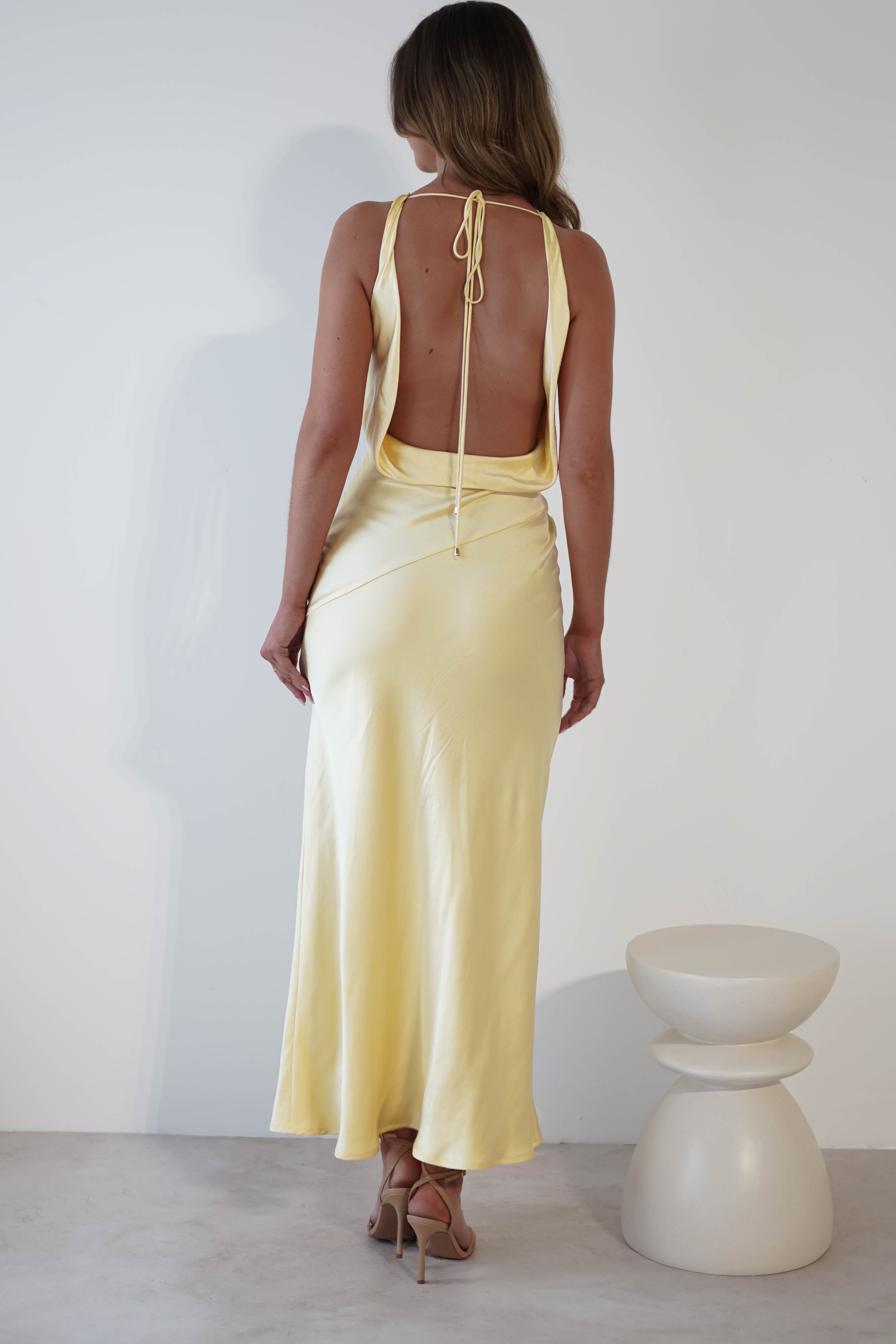 A woman in a draped open-back yellow satin dress with a tie at the neck stands facing a white wall. She wears beige high heels and her long hair cascades down her back. Next to her is a minimalist white stool with layered circular shapes.
