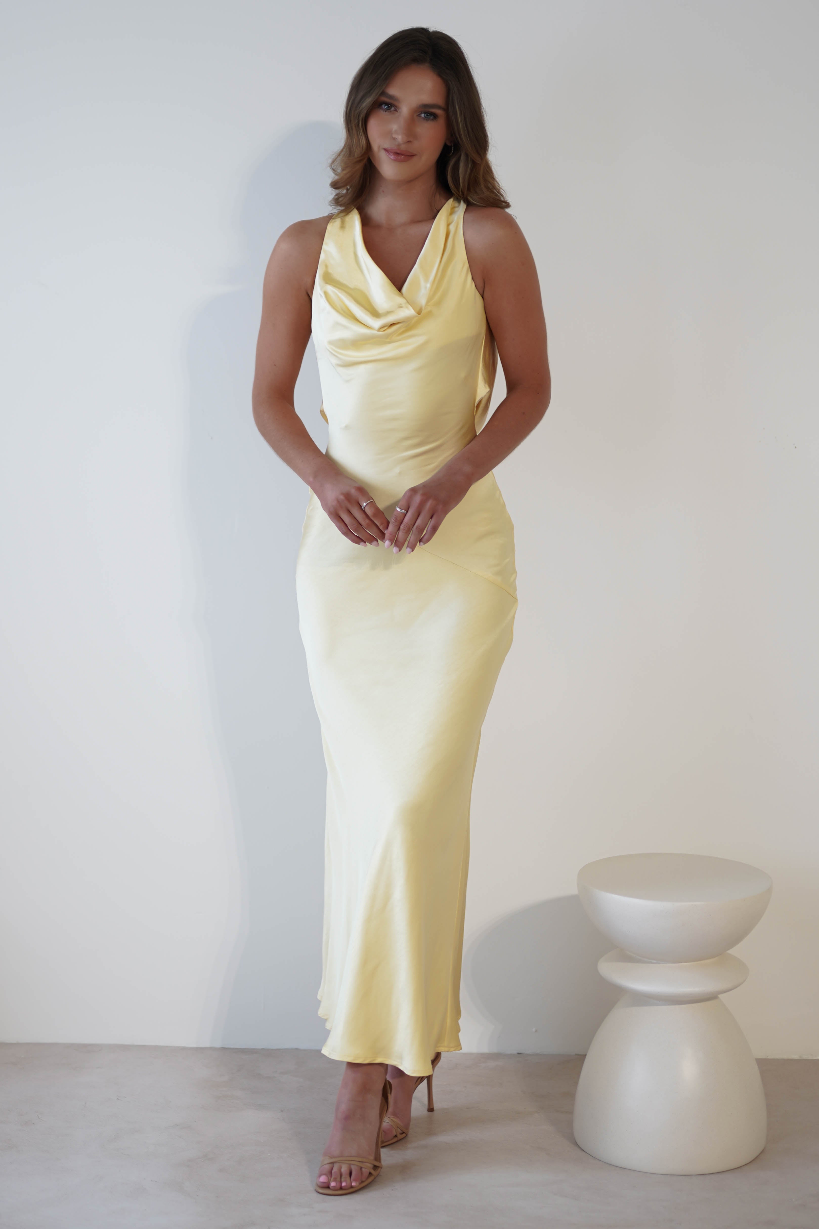 A woman in a long, sleeveless yellow dress with a cowl neckline and draped open-back stands against a plain white background beside a small white stool.