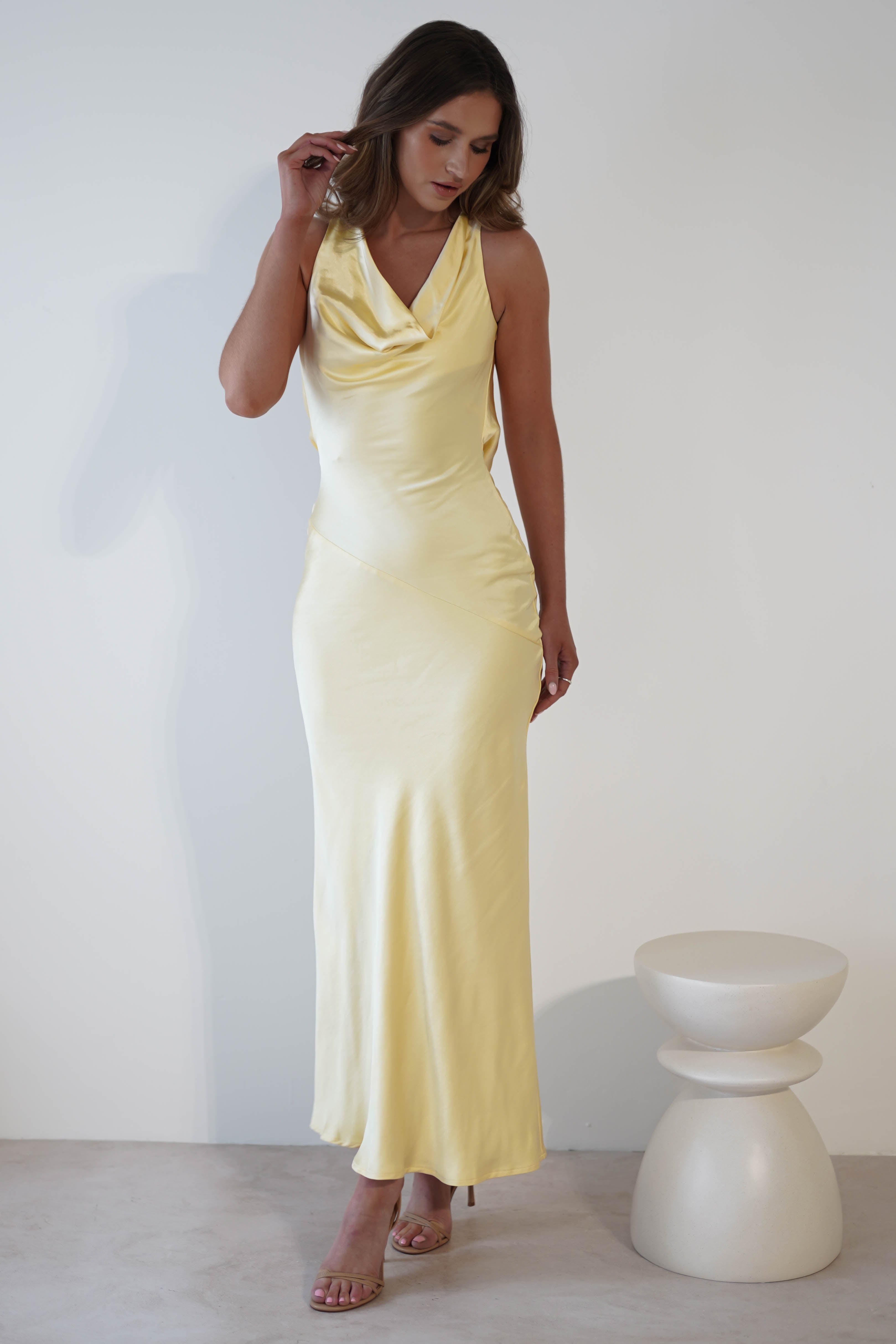 A person in a flowing, sleeveless light yellow dress with a draped open-back stands against a plain white background, touching their hair. A minimalist white stool is nearby. They wear heels, and the dress elegantly cascades to the floor.
