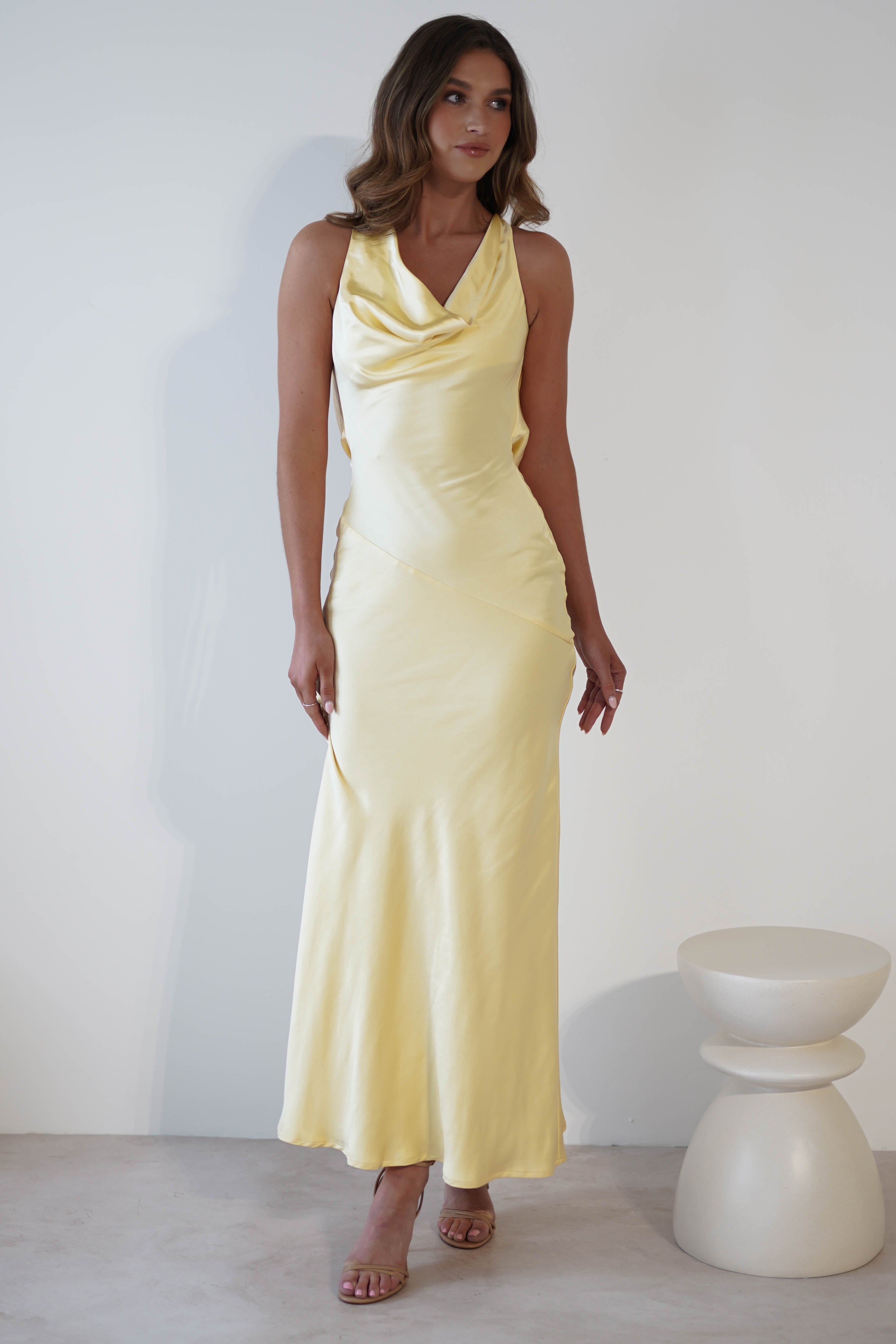 A person stands in a sleeveless, light yellow satin dress with a cowl neckline and an elegant draped open-back. They are wearing strappy heels and posing against a plain white wall. A small, round, white stool is visible to the right.