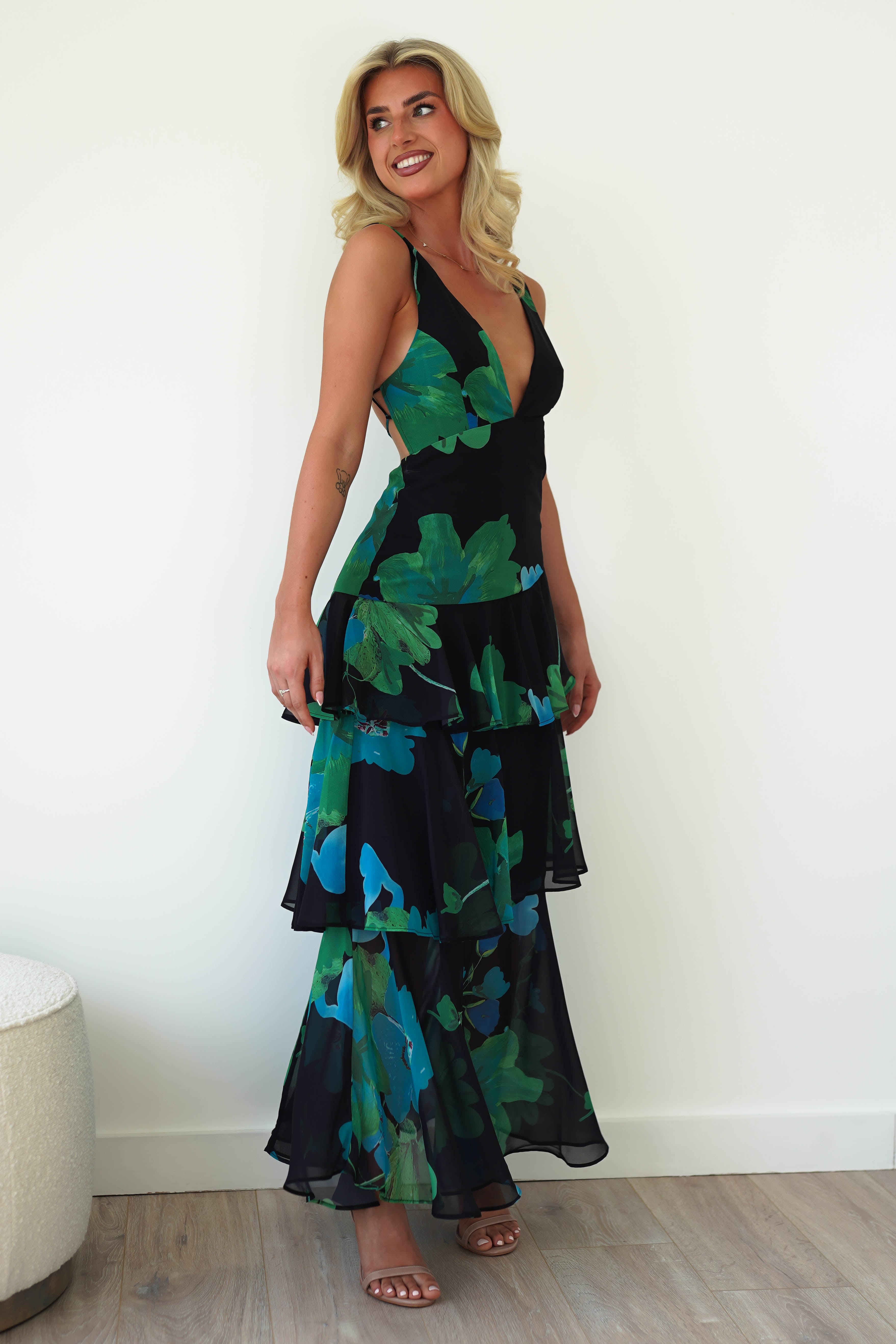 The woman, wearing a sleek black floral midi dress with green and blue designs, stands indoors. Her long, wavy blonde hair complements her joyous smile as she gazes sideways. The wooden floor and light-colored round stool add charm, making it perfect wedding guest attire in chiffon fabric.