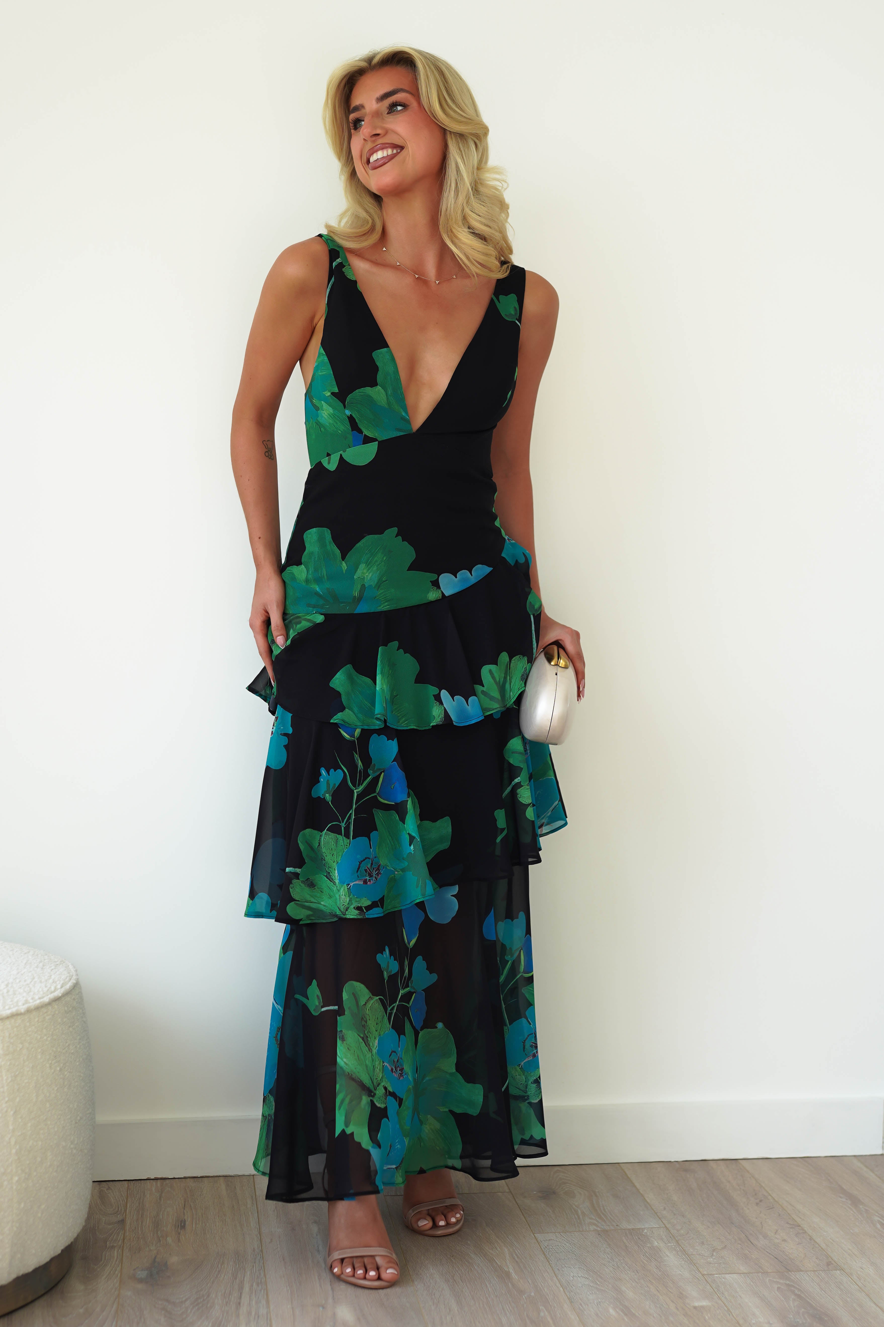A woman in a black floral midi with green patterns holds a clutch. The chiffon dress features a deep V-neck and tiered layers. With blonde hair, she smiles against a plain white wall, near light wood flooring and a small, round white stool—a perfect wedding guest outfit.