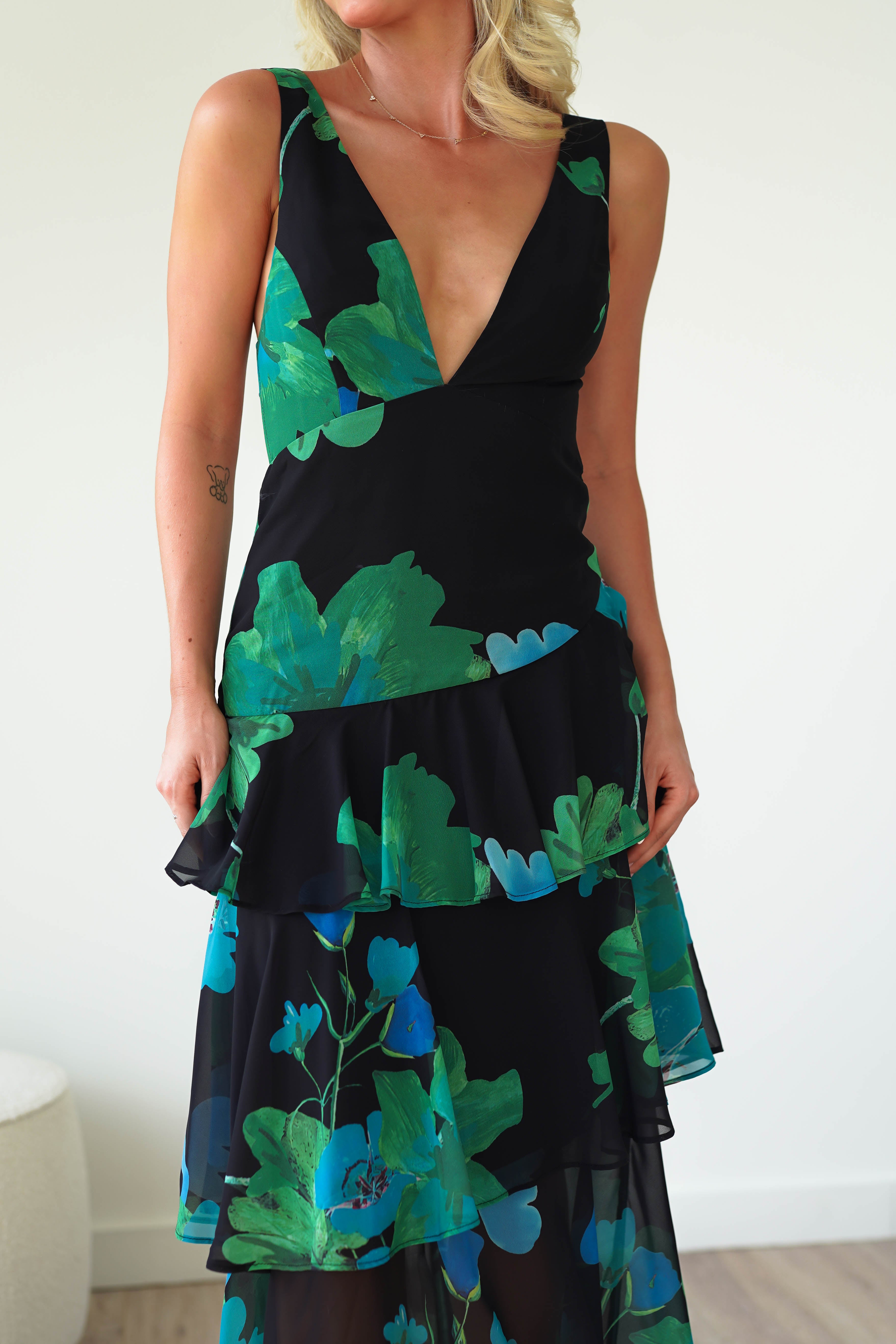 A woman elegantly dons a black floral midi dress, crafted from soft chiffon fabric, adorned with a deep V-neckline and green floral pattern. The dress showcases layered ruffles as she stands against a plain, light-colored background, perfect as wedding guest attire.