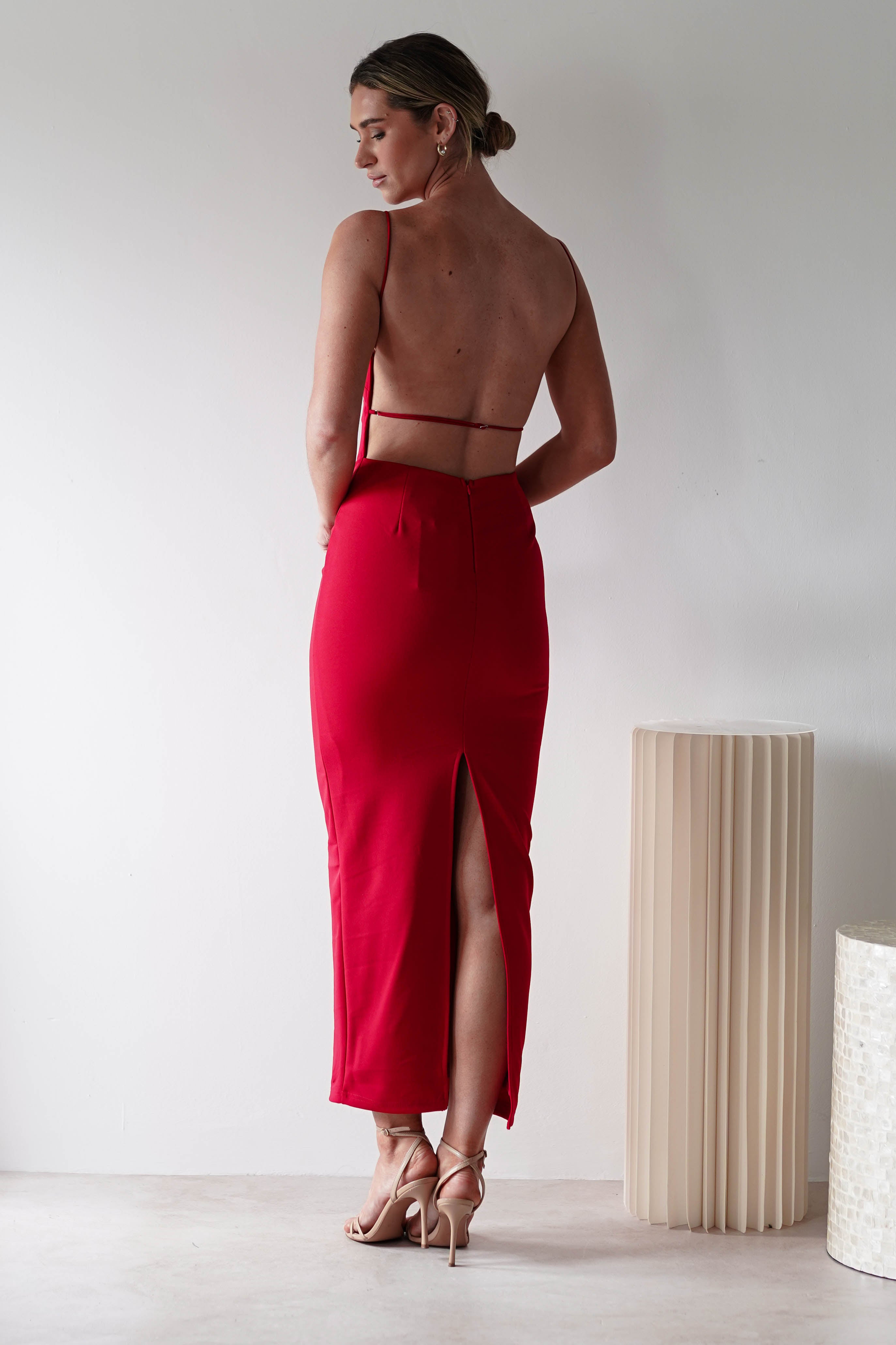 A woman in a red, backless, strappy dress with a slit and square neck stands in high heels, facing away. She stands near two modern, light-colored cylindrical lamps against a plain white wall—perfect for a stylish wedding guest look.