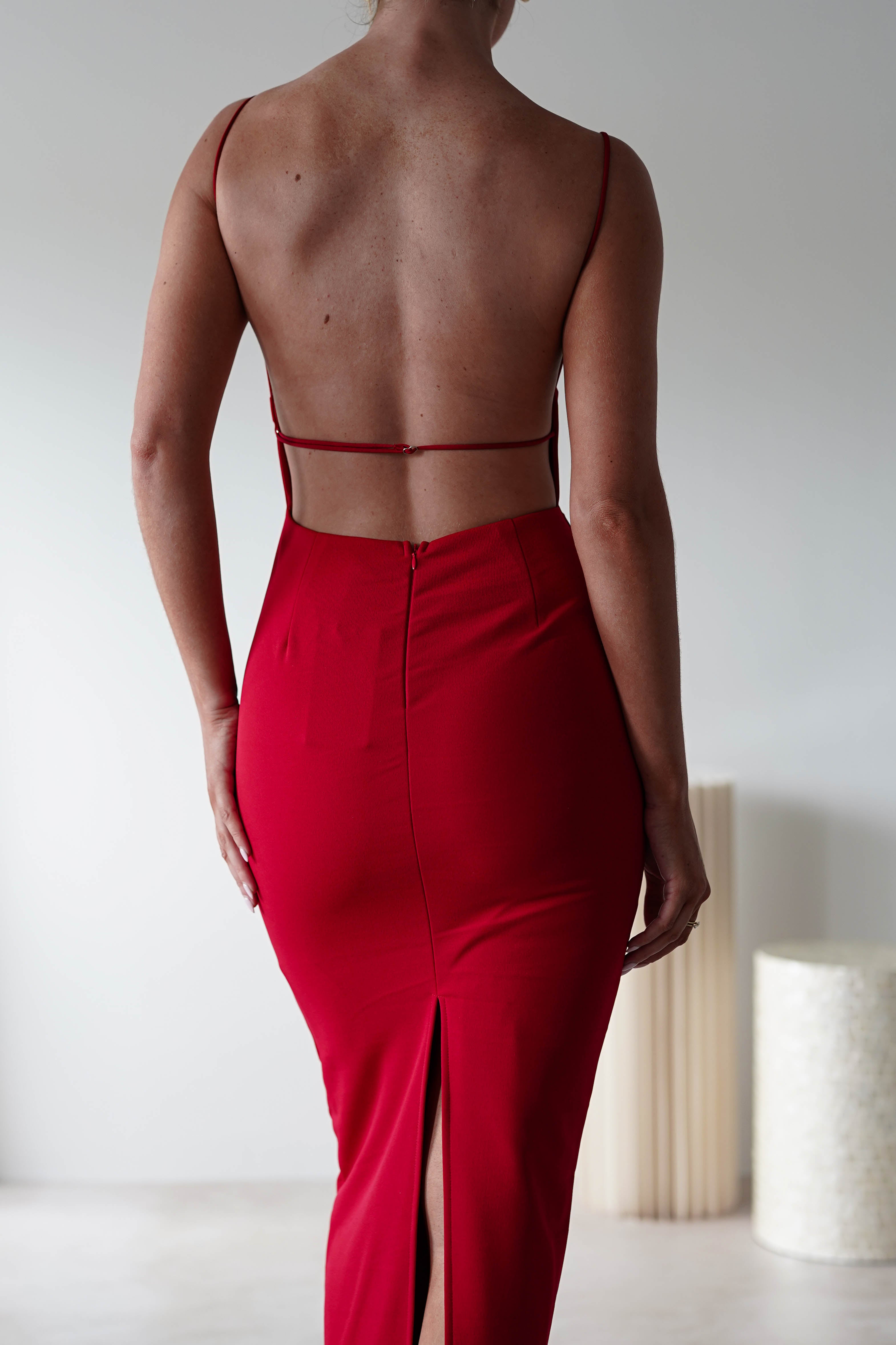 A person in a sleeveless, backless red dress with thin straps and a square neck stands facing away, revealing an open back and back slit. The stretchy material flatters the silhouette, perfect for a stylish wedding guest. The background is minimal and softly lit.