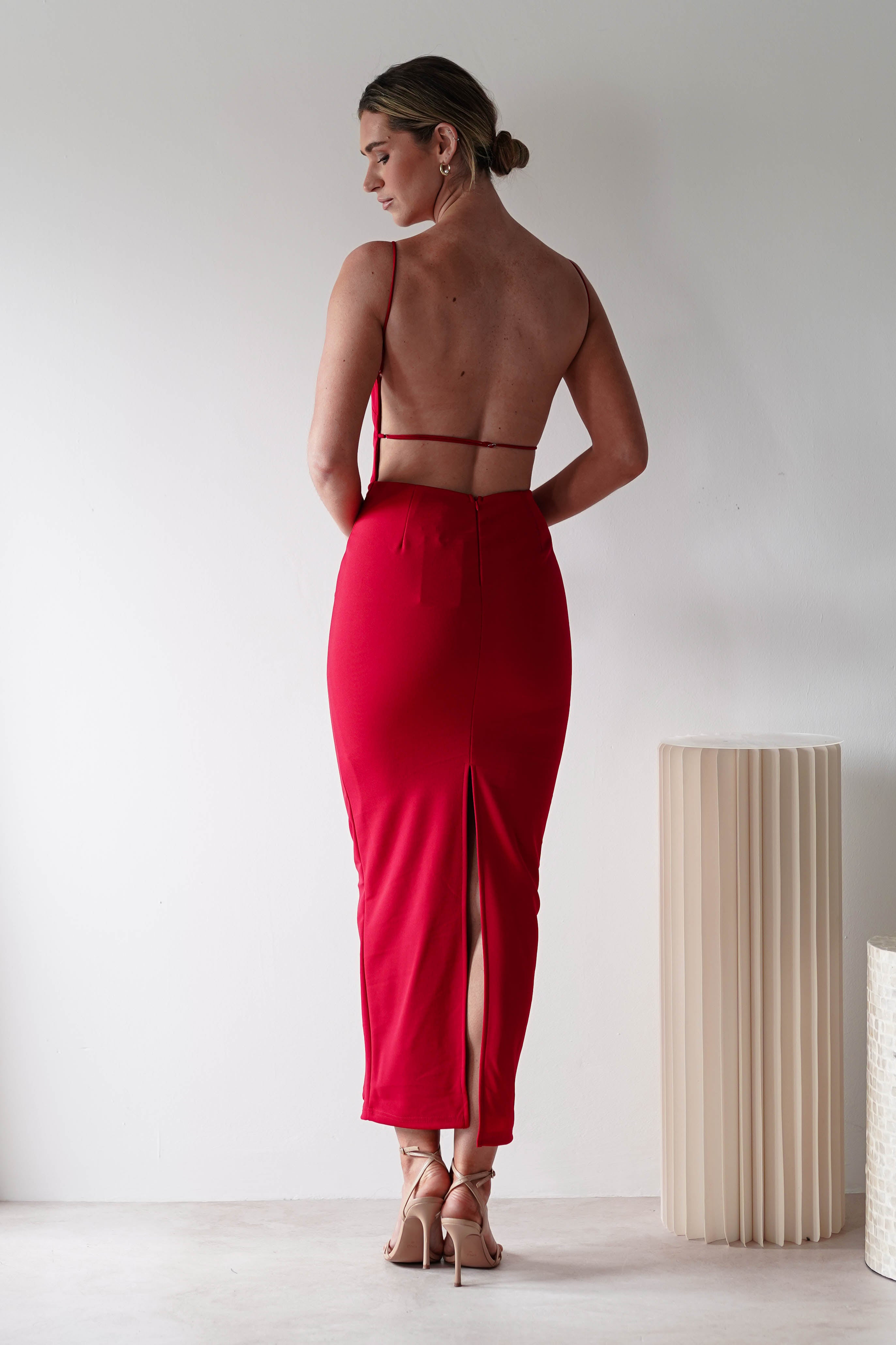 A woman stands facing away, wearing a fitted red backless dress with thin straps, a slit at the back, and crafted from stretchy material—perfect for a wedding guest. She pairs nude high heels with the look near a beige ribbed cylindrical side table.