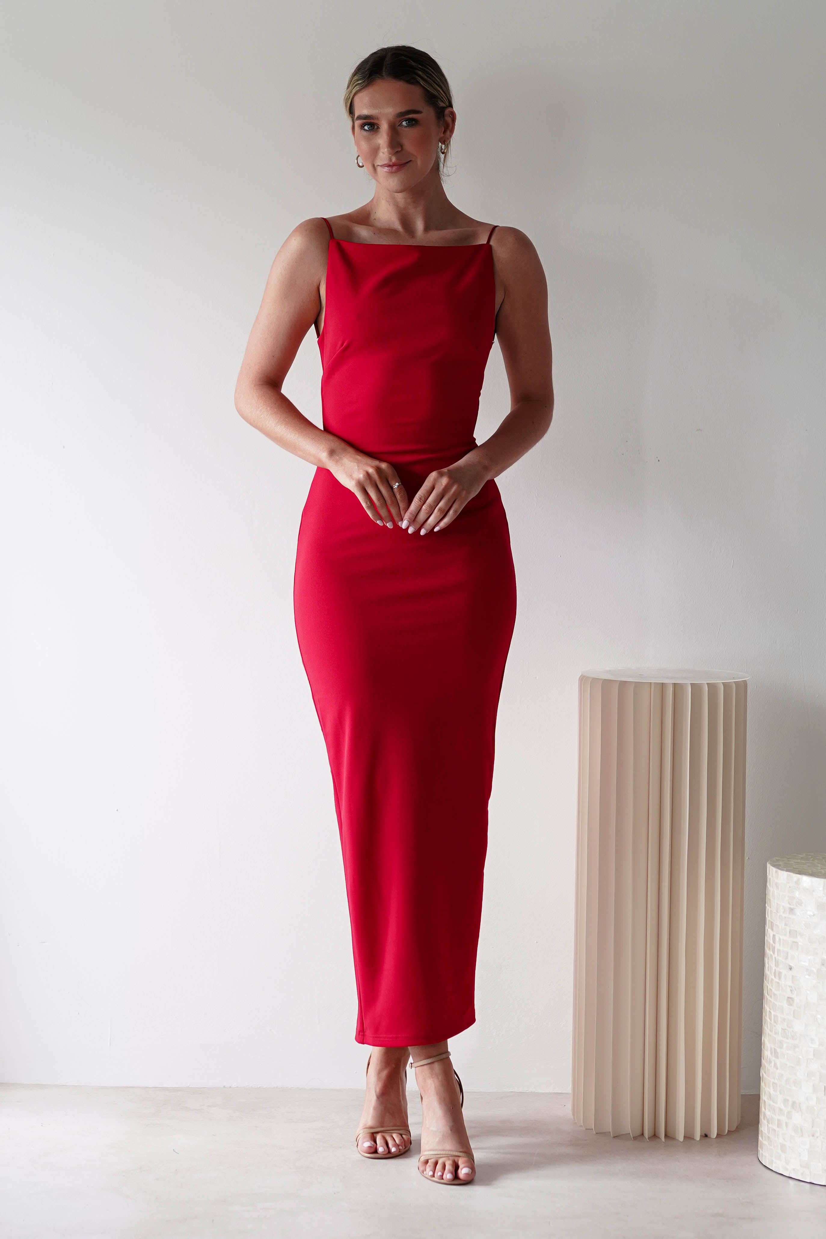 A woman stands indoors against a plain white wall, wearing a fitted, sleeveless red dress with a square neck and nude heels—an ideal look for a wedding guest. She clasps her hands and smiles slightly; a modern cylindrical lamp is by her side.