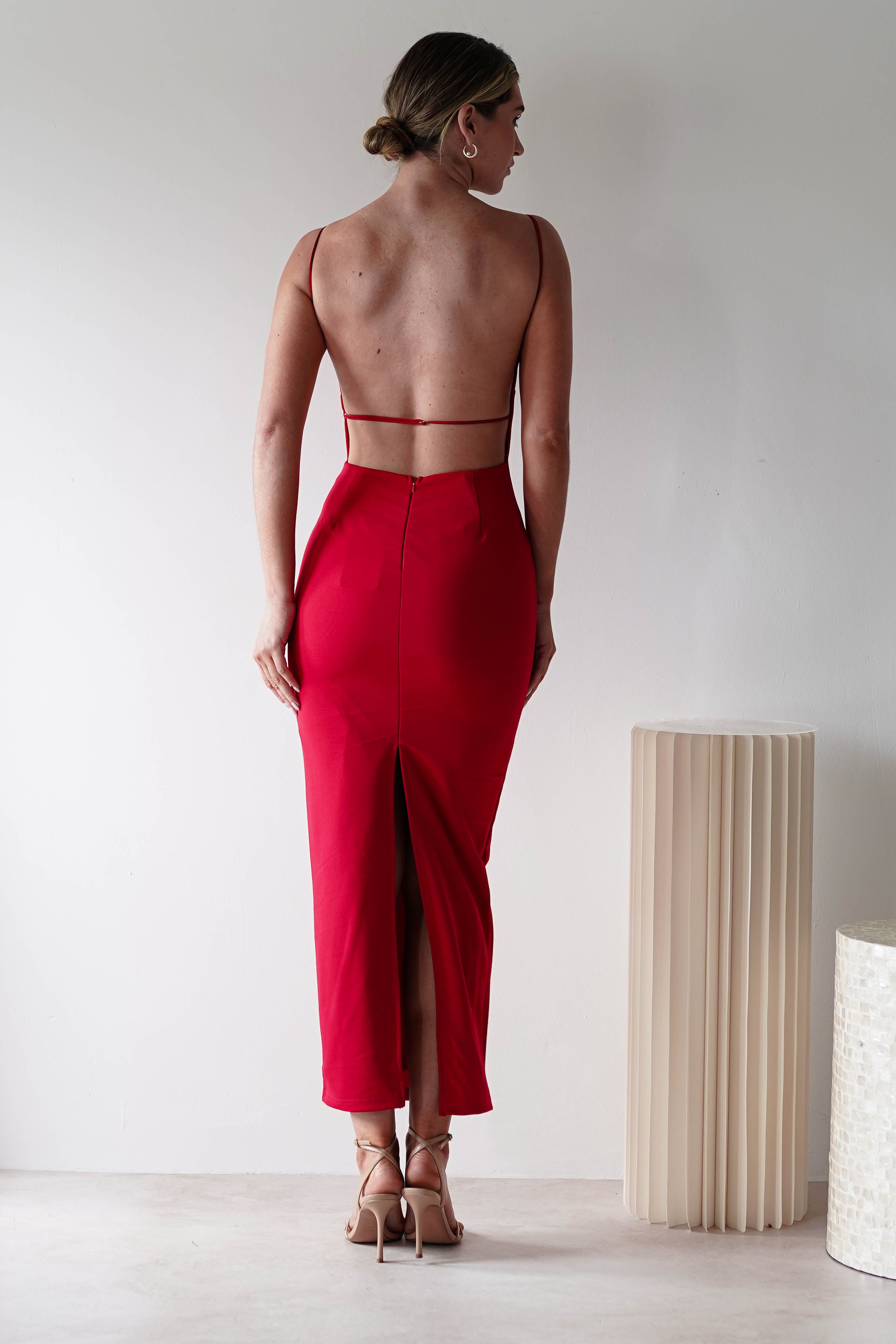 A woman stands facing away, wearing a fitted red dress with a square neck, backless design, spaghetti straps, and a back slit. Her hair is neatly tied up and she pairs nude heels with the look. Minimalist decor in neutral tones surrounds her.
