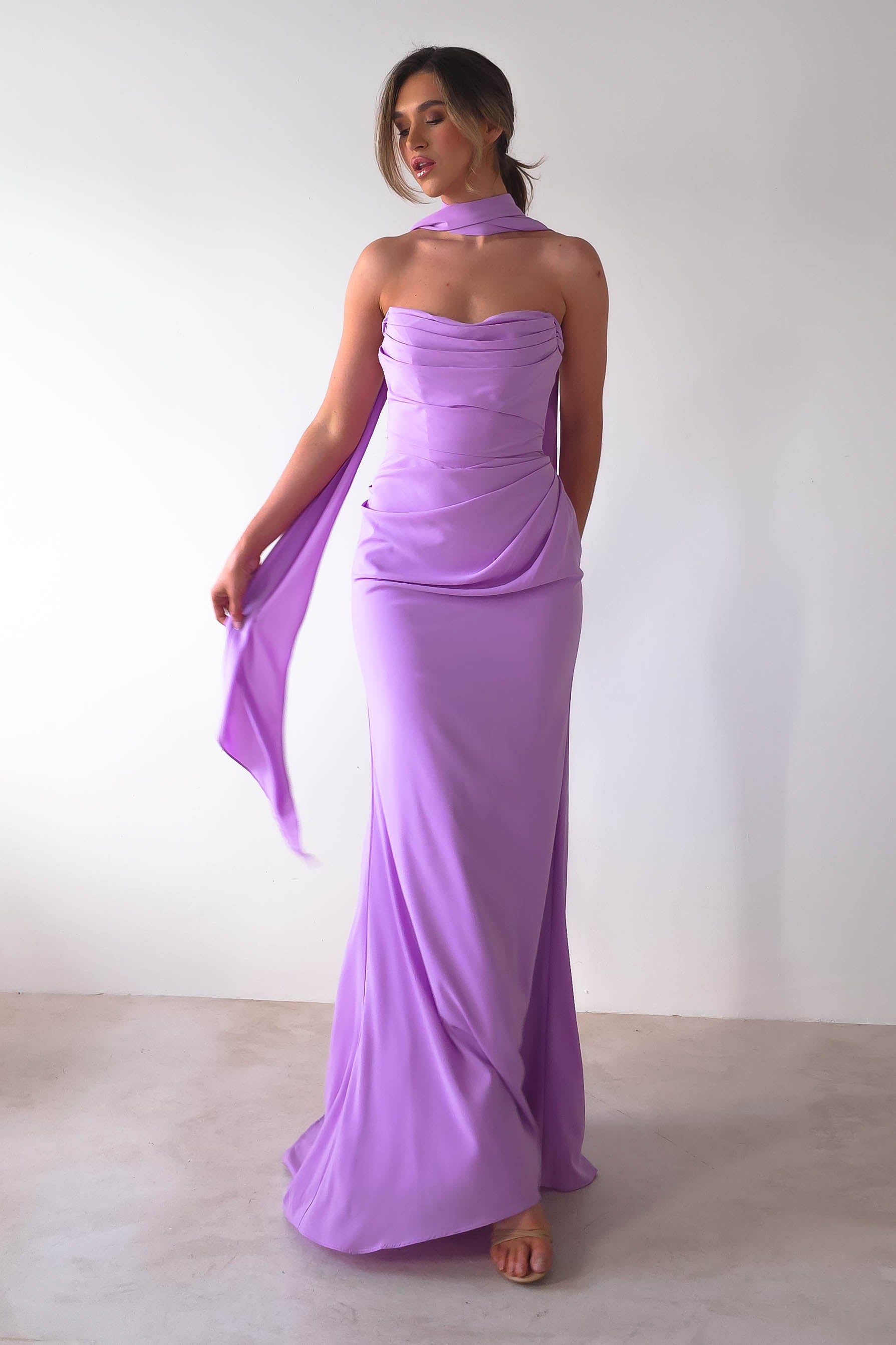 A woman stands against a plain white background, elegantly wearing the strapless design of the Lisa Multiway Chiffon Gown in lavender with a matching scarf. Her hair is styled up as she poses gracefully, barefoot on the floor-length hem.