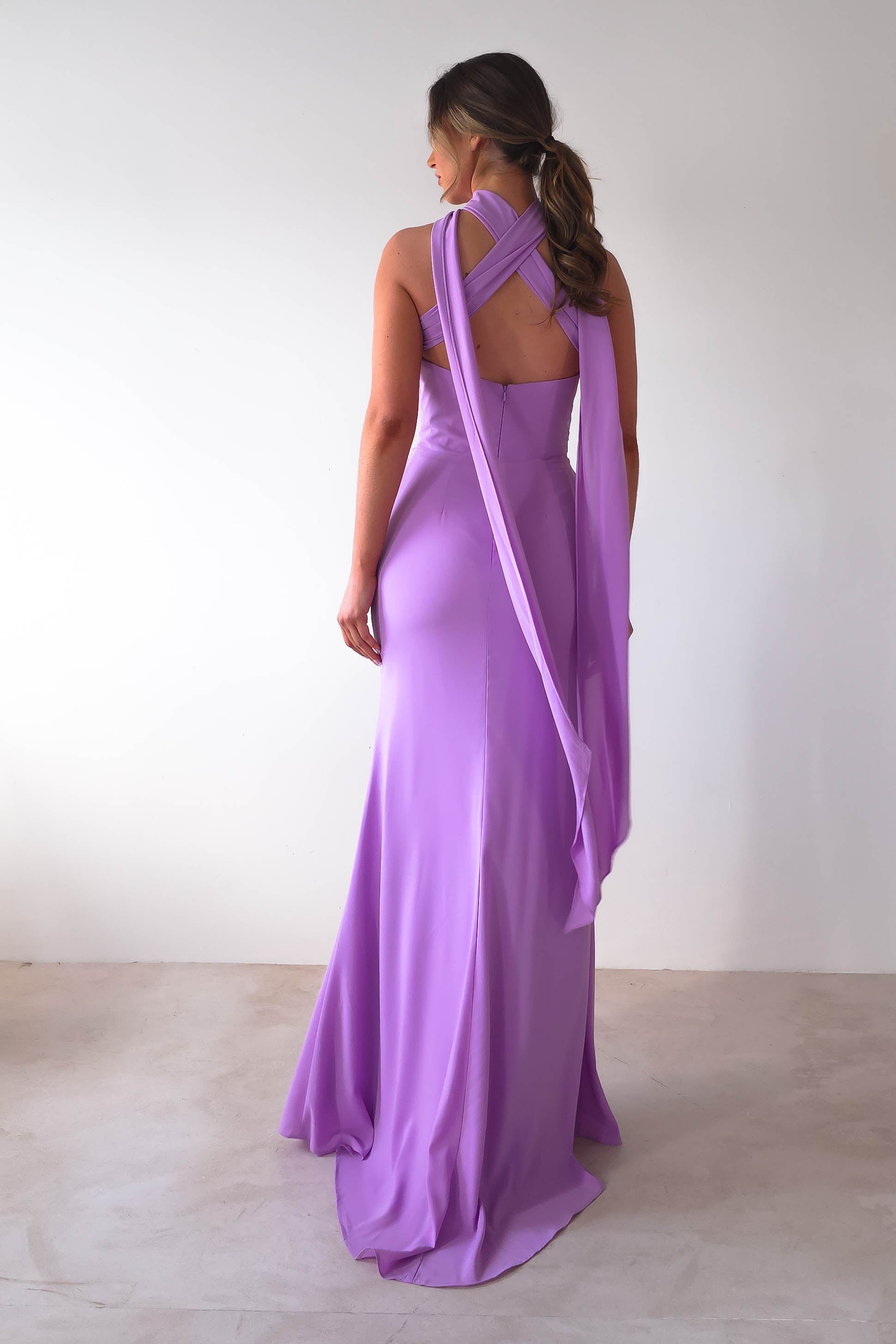 A woman stands with her back to the camera, wearing the elegant Lisa Multiway Chiffon Gown in lavender. Its versatile ties create crossed straps and a flowing train. Her hair is styled in loose waves as she poses against a plain white backdrop, exuding timeless grace.