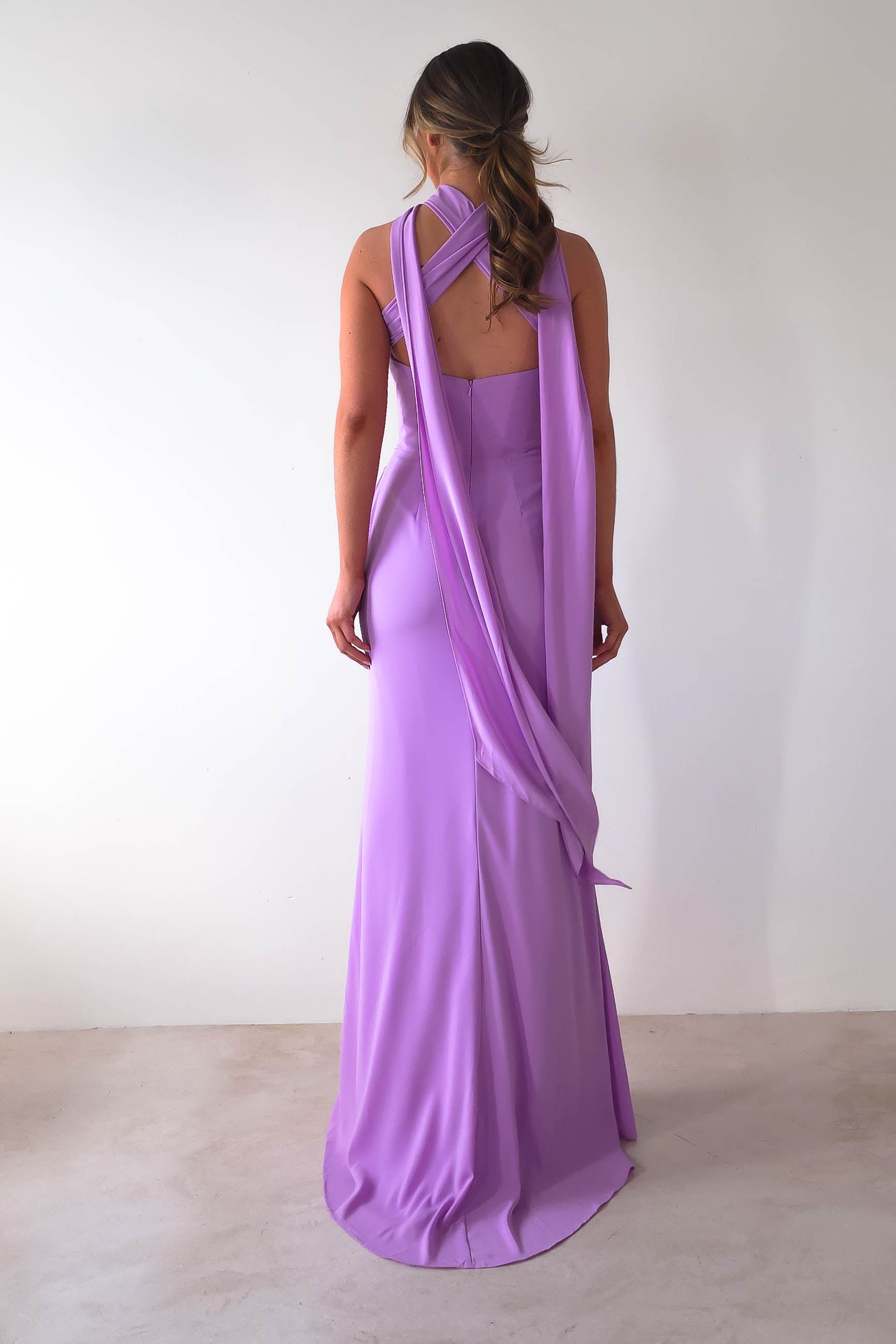 Rear view of a person in a lavender Lisa Multiway Chiffon Gown, showcasing a unique draped back design and crisscross straps. The floor-length gown flows smoothly against the plain white wall, while their hair cascades in loose waves, enhancing the gowns elegant versatility.