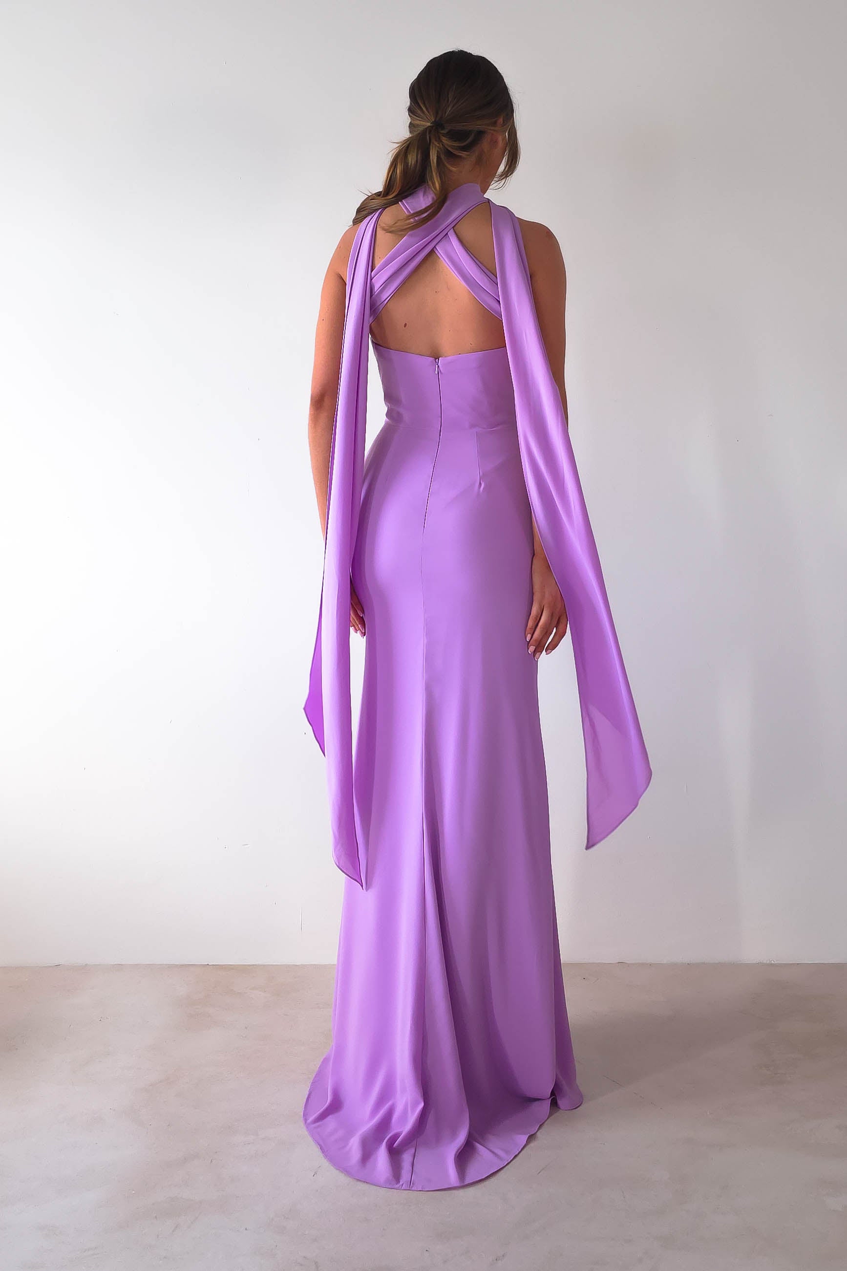 A woman stands facing away, dressed in the Lisa Multiway Chiffon Gown—a floor-length, lavender masterpiece with a crisscross halter neck. Its versatile ties cascade down her back, offering a fitted silhouette against the plain white wall.