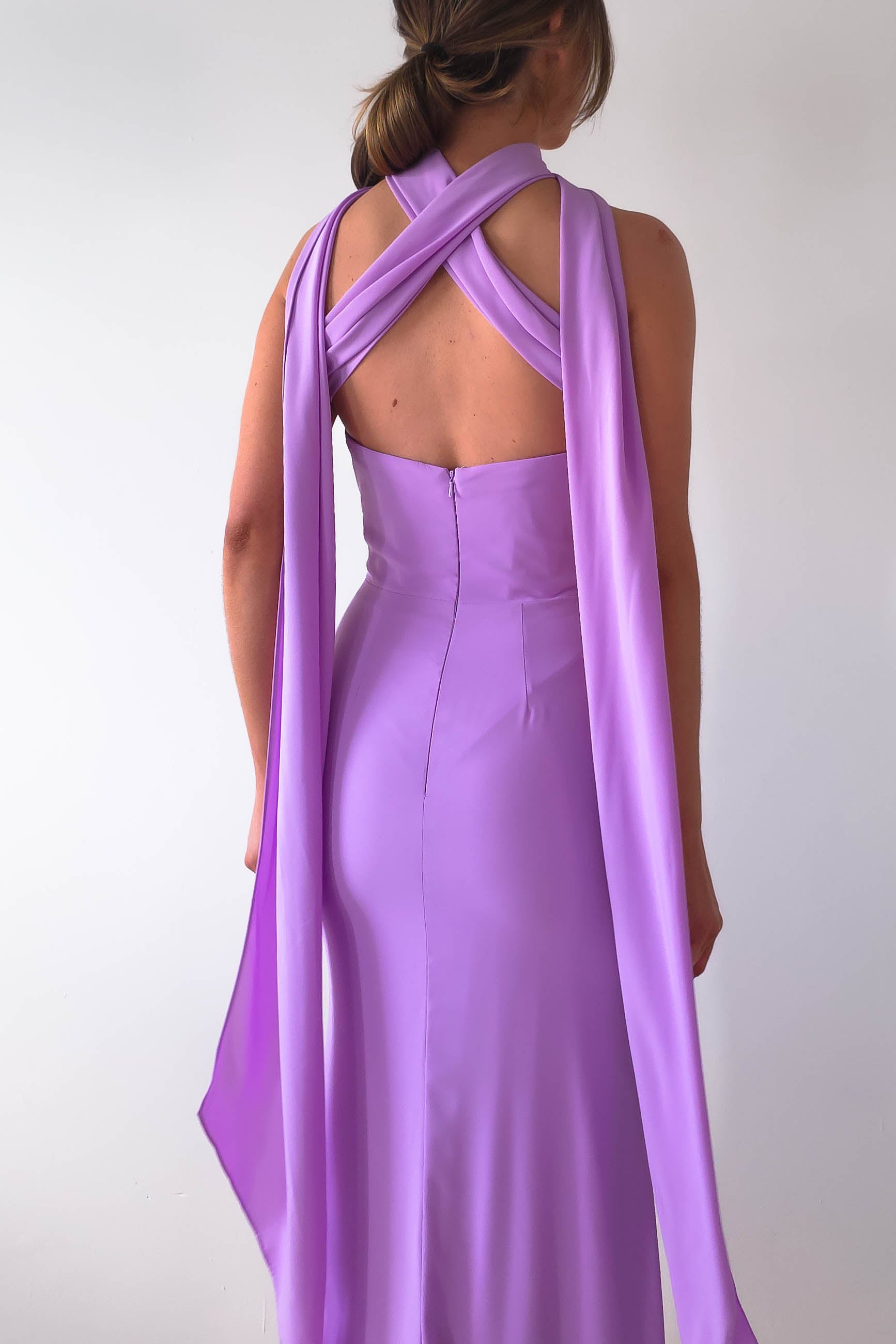 A person wearing the Lisa Multiway Chiffon Gown, featuring a floor-length, sleeveless lavender design with elegant crisscross back details. The versatile ties add sophistication as they drape over the shoulders against a plain white backdrop.