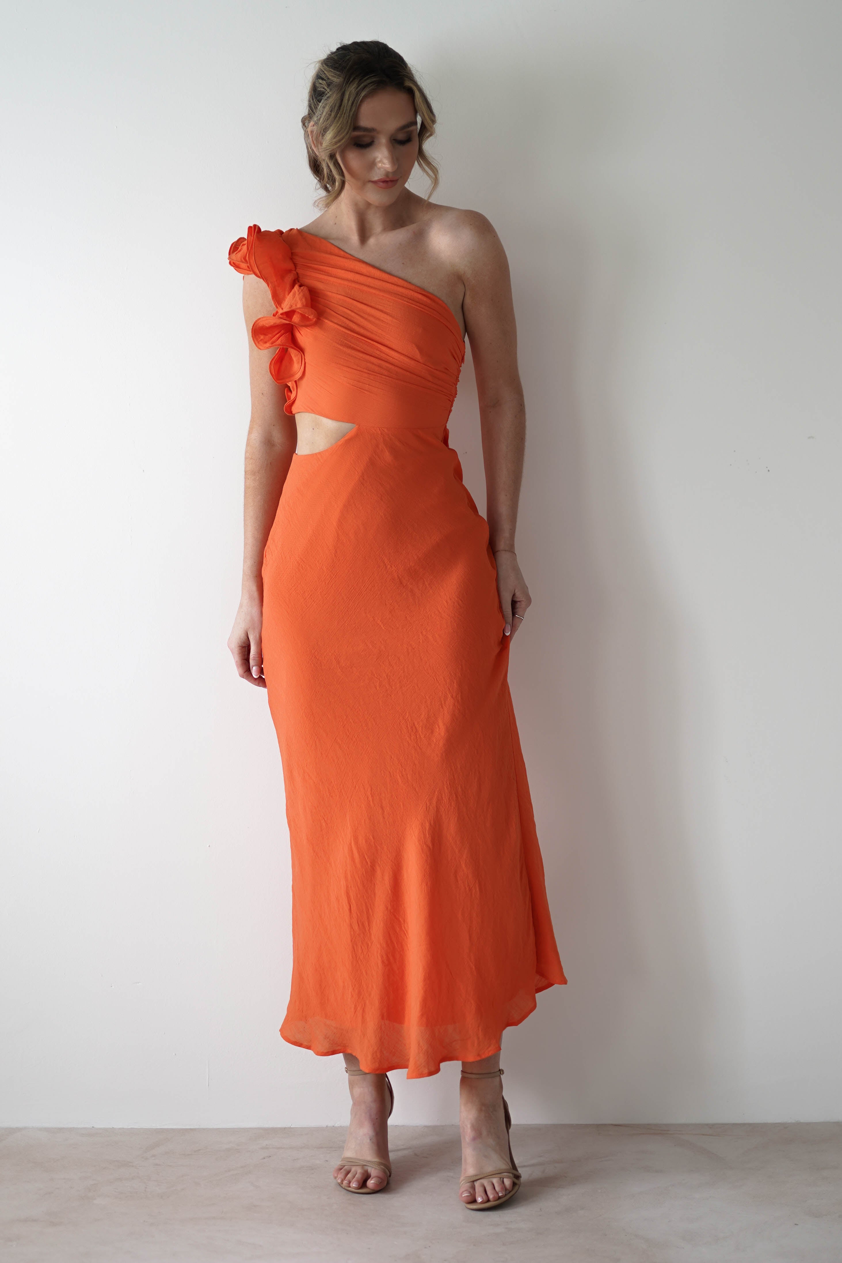 A woman stands against a plain light wall, wearing a bright orange one-shoulder ruffle maxi with an ankle-length hem and midriff cut out, perfect for special occasions. She has wavy hair and pairs the look with nude strappy heels.