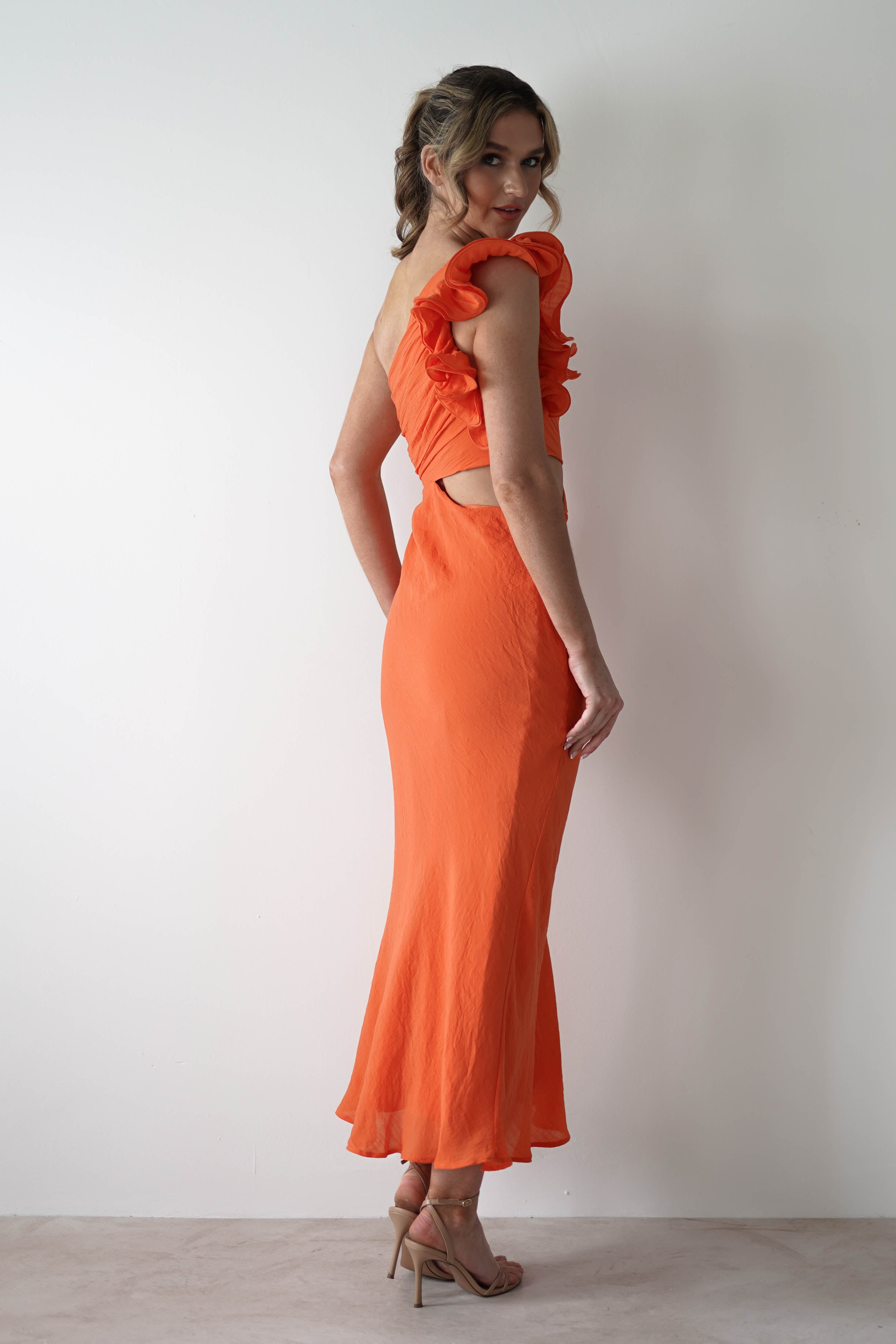 Woman standing sideways against a white wall, wearing a one shoulder ruffle maxi dress with midriff cut outs and beige high-heeled sandals. She looks over her shoulder and smiles slightly—perfect for special occasions.