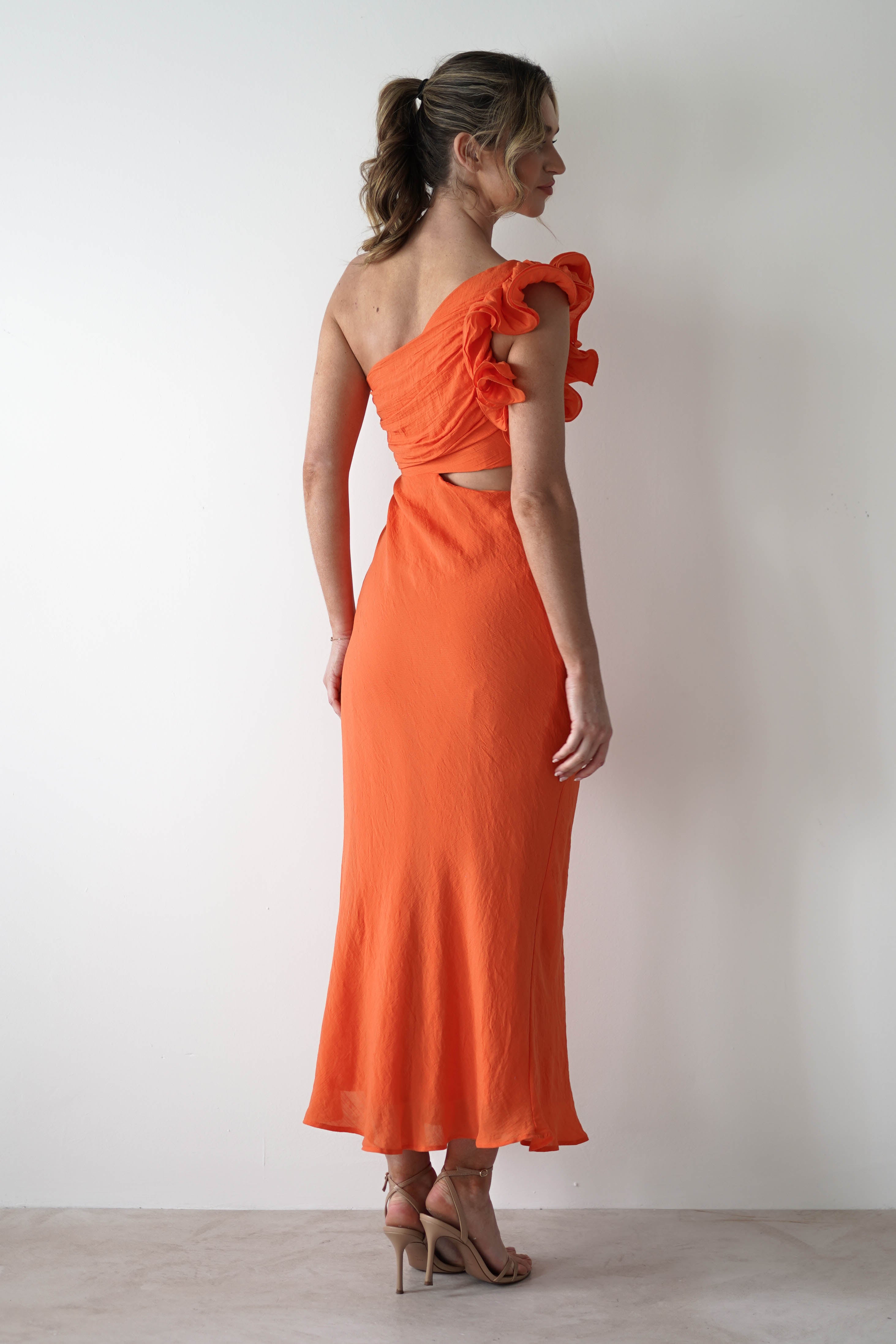 A woman stands facing away, wearing an orange one shoulder ruffle maxi dress with midriff cut outs. Her hair is tied back in a ponytail, and she pairs beige high-heeled sandals with the look—perfect for special occasions against a light background.