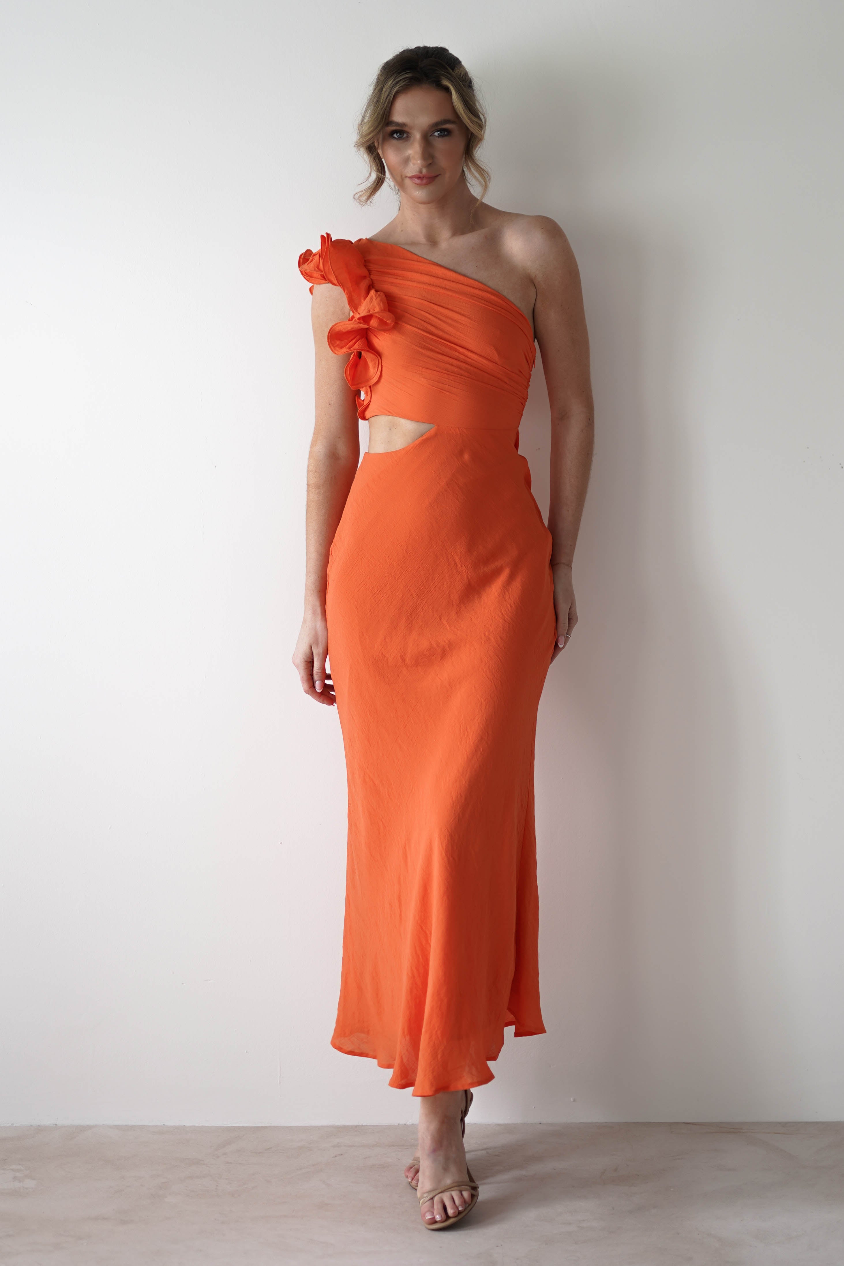 A woman stands against a plain white wall in a one shoulder ruffle maxi dress with midriff cut outs—perfect for special occasions. Her light hair is styled up, and she completes the look with nude heels.