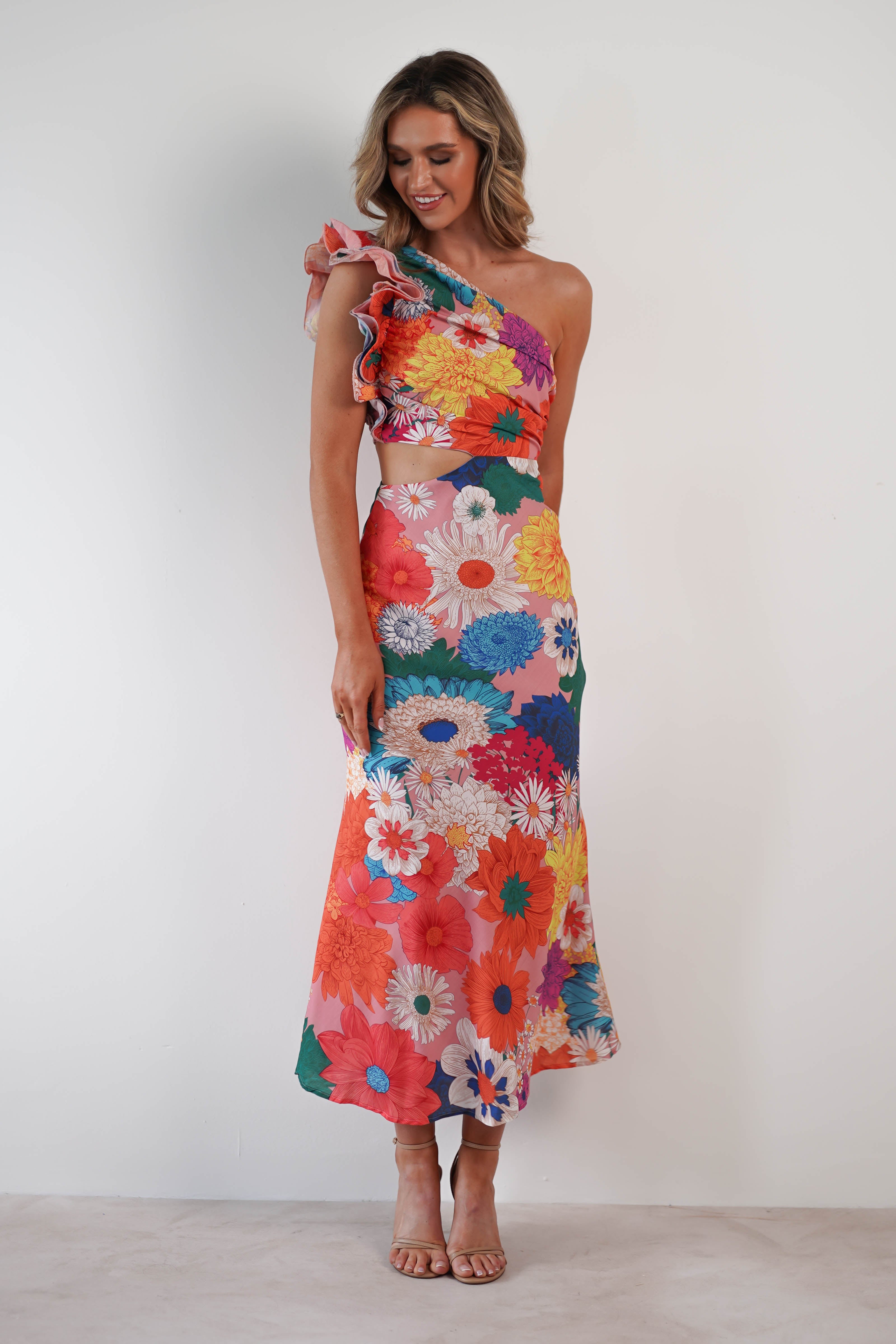 Woman in a colorful, one-shoulder ruffle maxi with a vibrant floral pattern and textured fabric smiles while standing against a plain background. She has wavy hair and wears strappy high heels.