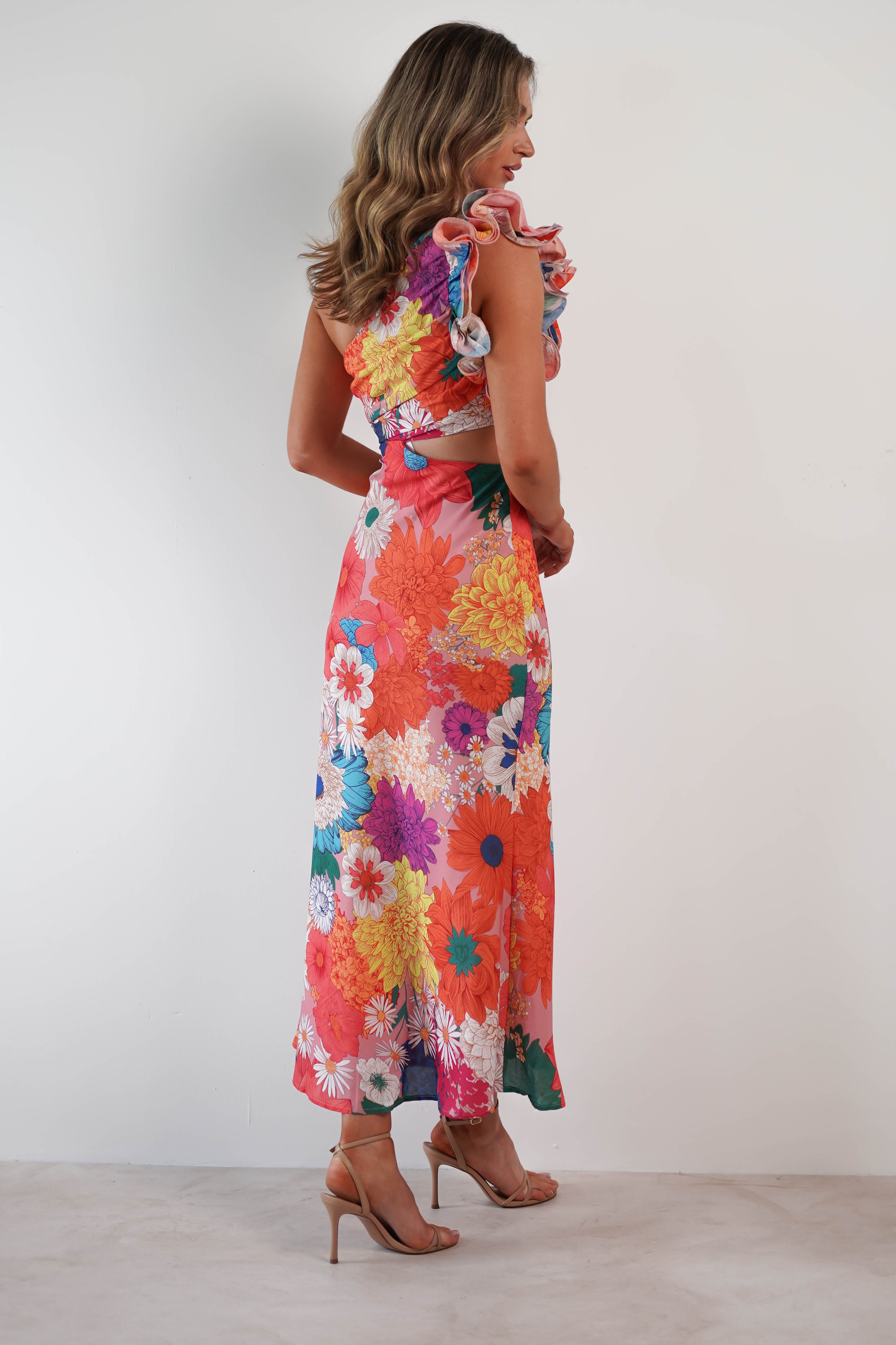 A person stands against a white backdrop, showcasing a one-shoulder ruffle maxi dress adorned with colorful, floral patterns. The textured fabric flaunts shades of red, pink, orange, and white flowers. Nude high-heeled sandals complement their look while long, wavy hair cascades down.