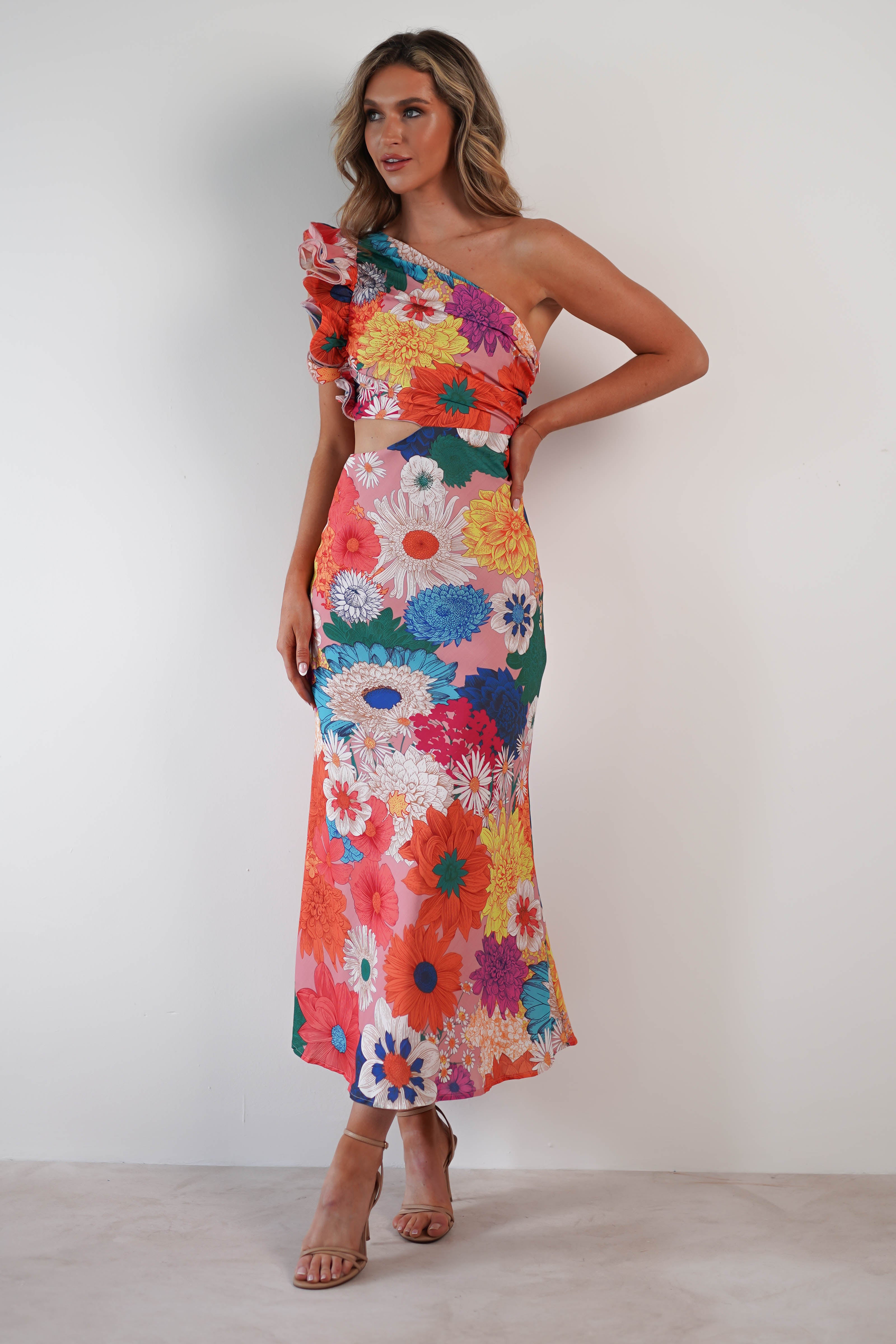 A person stands against a white background wearing a vibrant, one-shoulder ruffle maxi dress with large floral patterns in red, blue, and yellow. Featuring elegant midriff cut-outs, the dress complements their long, wavy hair as they pose confidently in strappy heels.