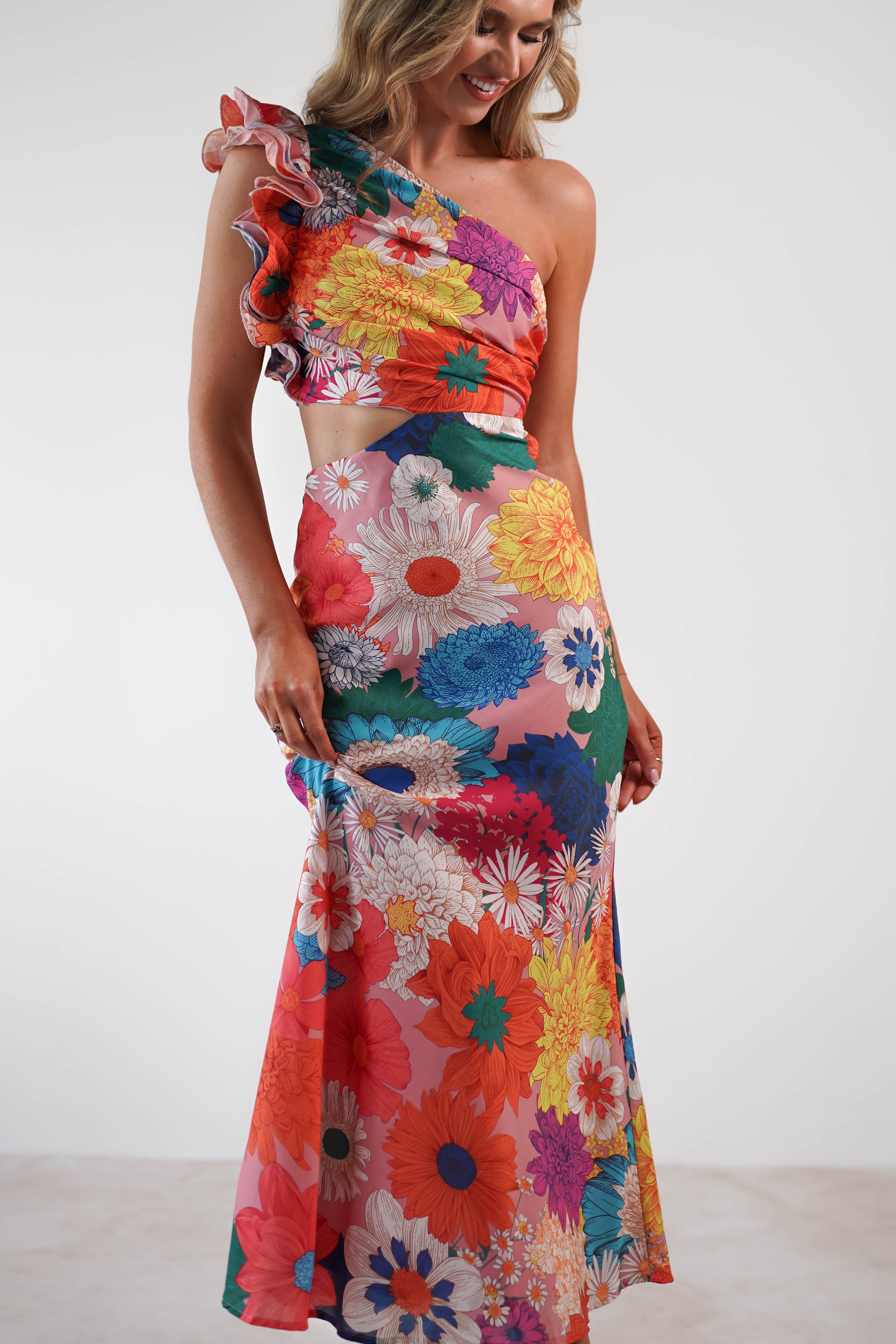 A woman wearing a colorful, one-shoulder floral dress with textured fabric, featuring a mix of large, vibrant flowers. The dress includes ruffled detail on the shoulder and gracefully falls as a maxi to the floor. She smiles warmly while standing against a plain background.