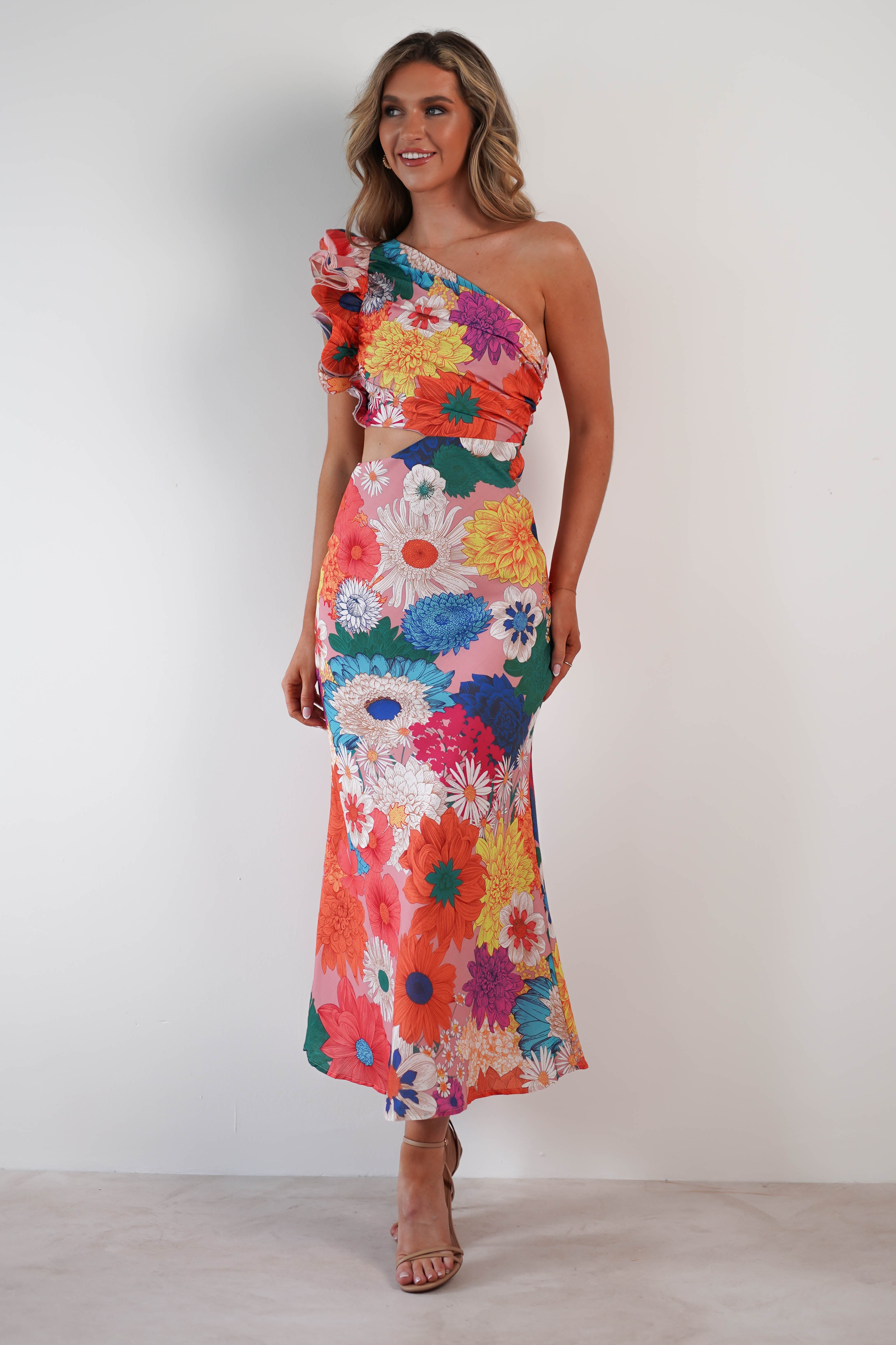 A person stands against a plain background, wearing a vibrant, floral one-shoulder ruffle maxi dress. The textured fabric showcases large flowers and an asymmetrical ruffle on the shoulder. They have long hair and are smiling.