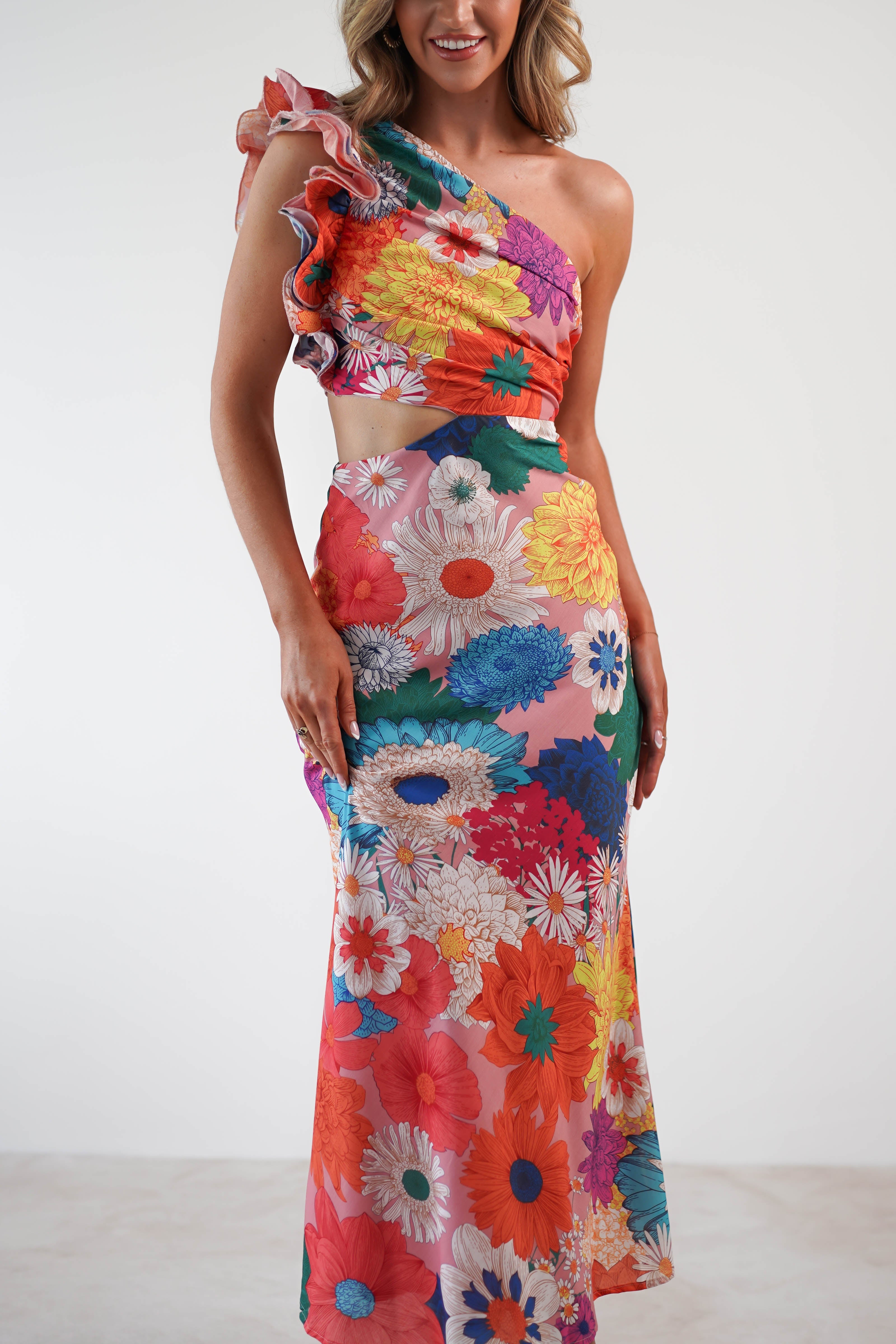 A person wearing a vibrant, one-shoulder ruffle maxi dress adorned with colorful floral patterns and large, bold blooms in red, blue, orange, pink, and white. The textured fabric enhances the elegant ruffled detailing on the shoulder as it extends to ankle length.