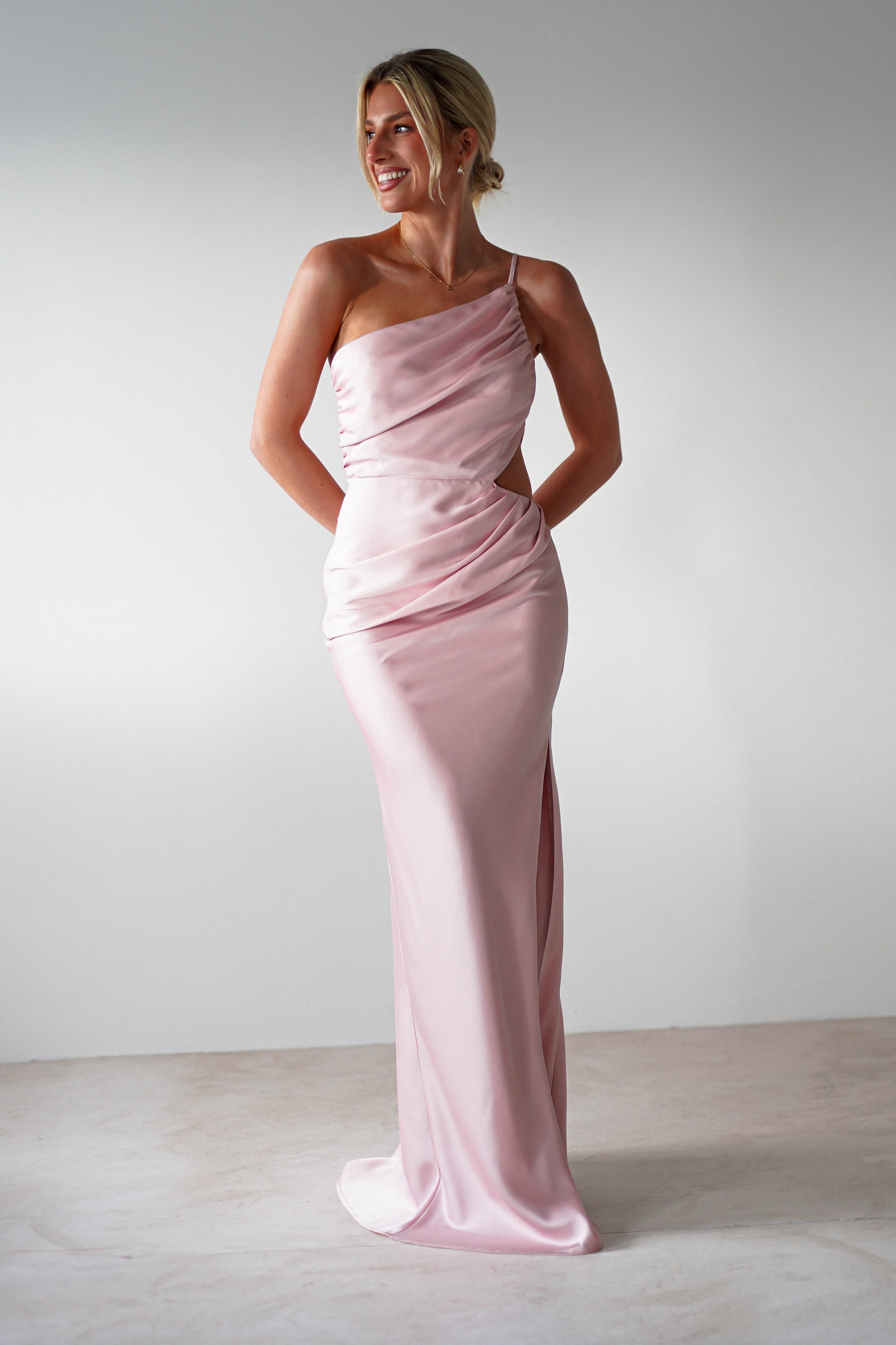 A woman stands smiling, wearing a long, light pink one-shoulder satin maxi that makes a stunning bridesmaids dress. Her hands are behind her back as she poses against a plain white background.