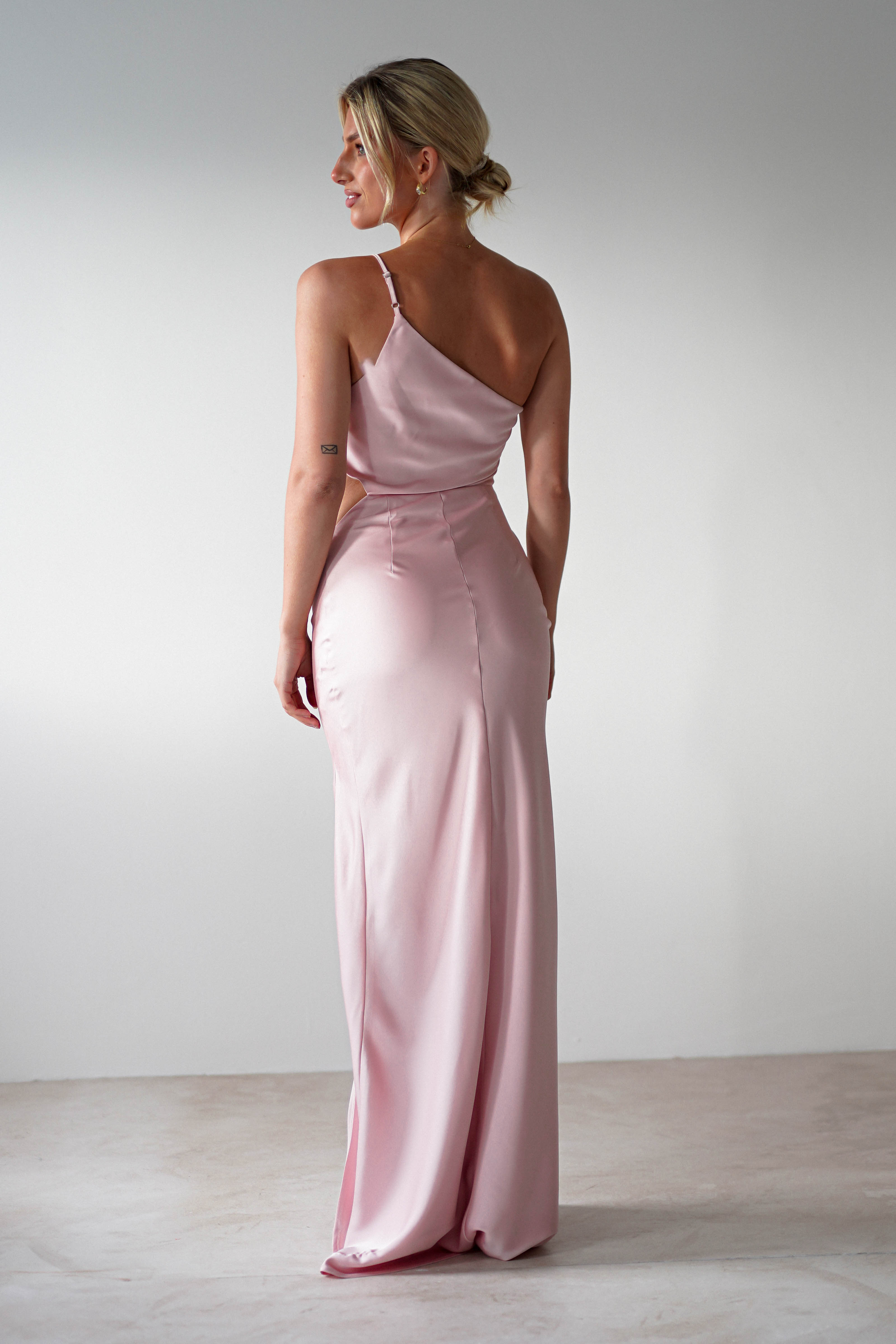 A woman stands facing away, wearing a one-shoulder satin maxi in light pink—a chic choice for a wedding guest dress. Her blonde hair is styled in a low bun, and she is in a minimal, neutral-colored room.
