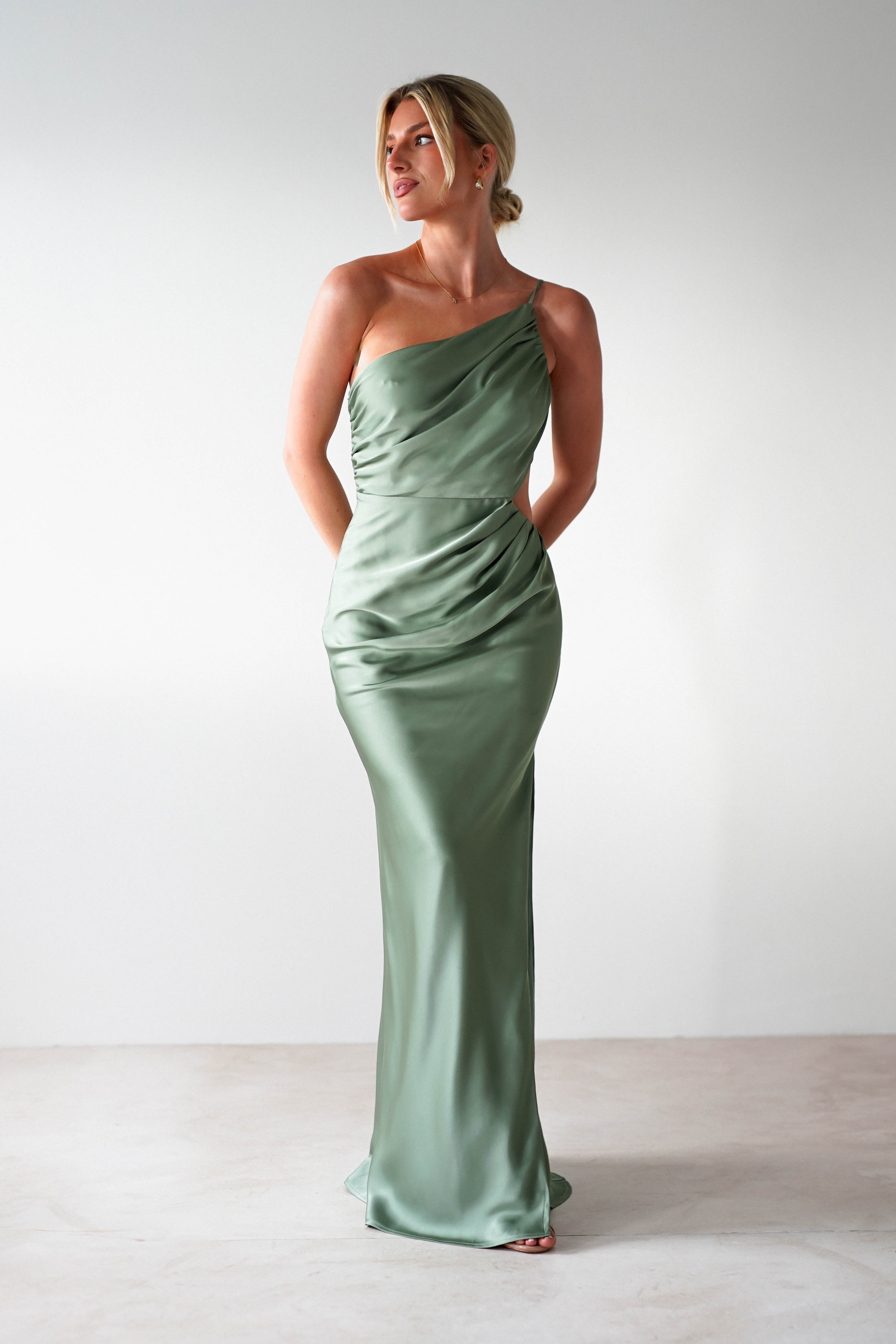 A person wearing a sleek, light green satin maxi dress stands with hands behind their back. The floor-length gown has a smooth texture and draped design. The background is plain white, and their hair is styled in an elegant updo, perfect for a wedding guest look.