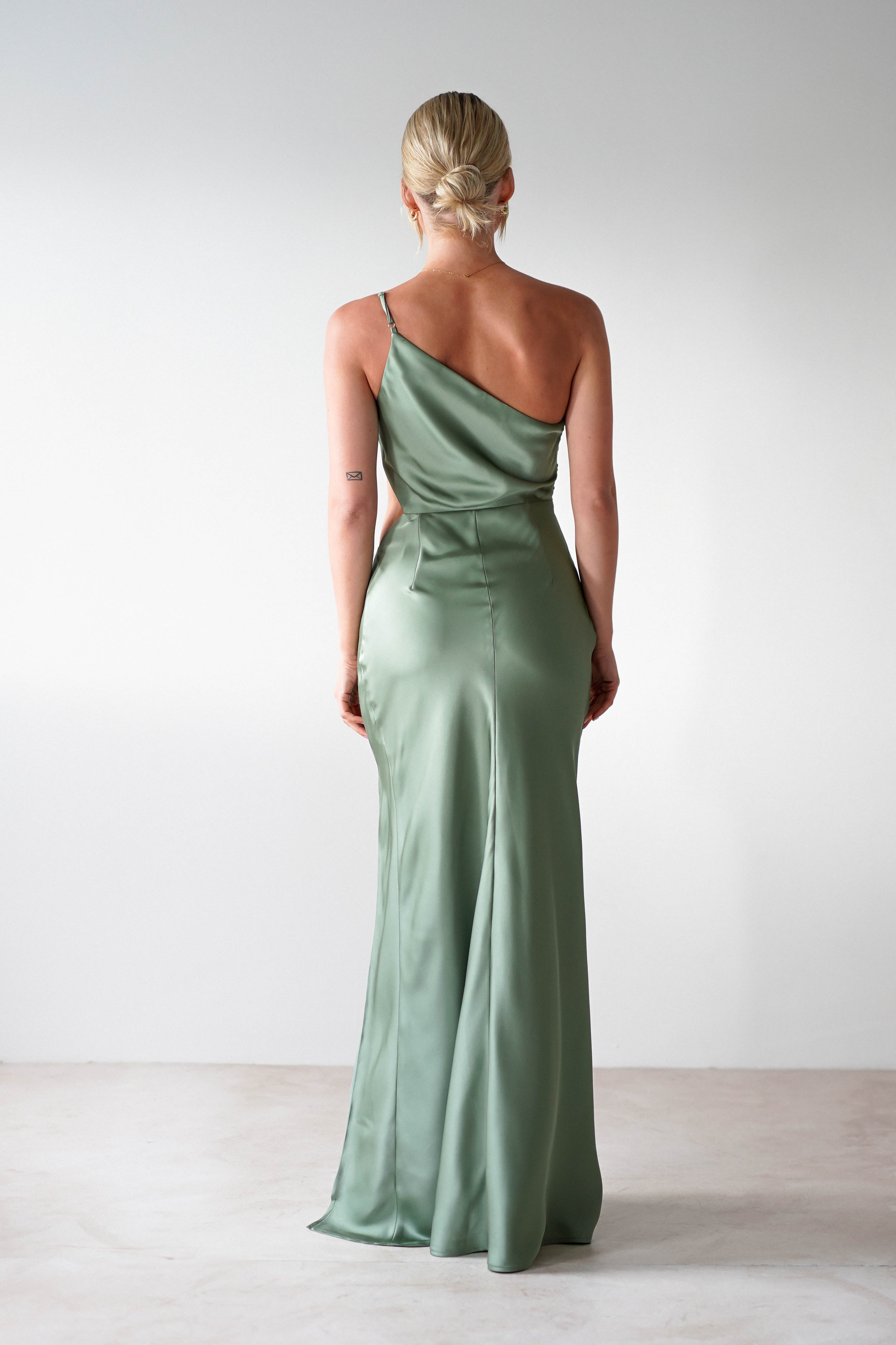 A person with blonde hair in an elegant updo is seen from behind, wearing a light green satin maxi dress with one shoulder strap. They stand on a light-colored floor against a plain white wall, perfectly embodying the grace of a wedding guest dress.