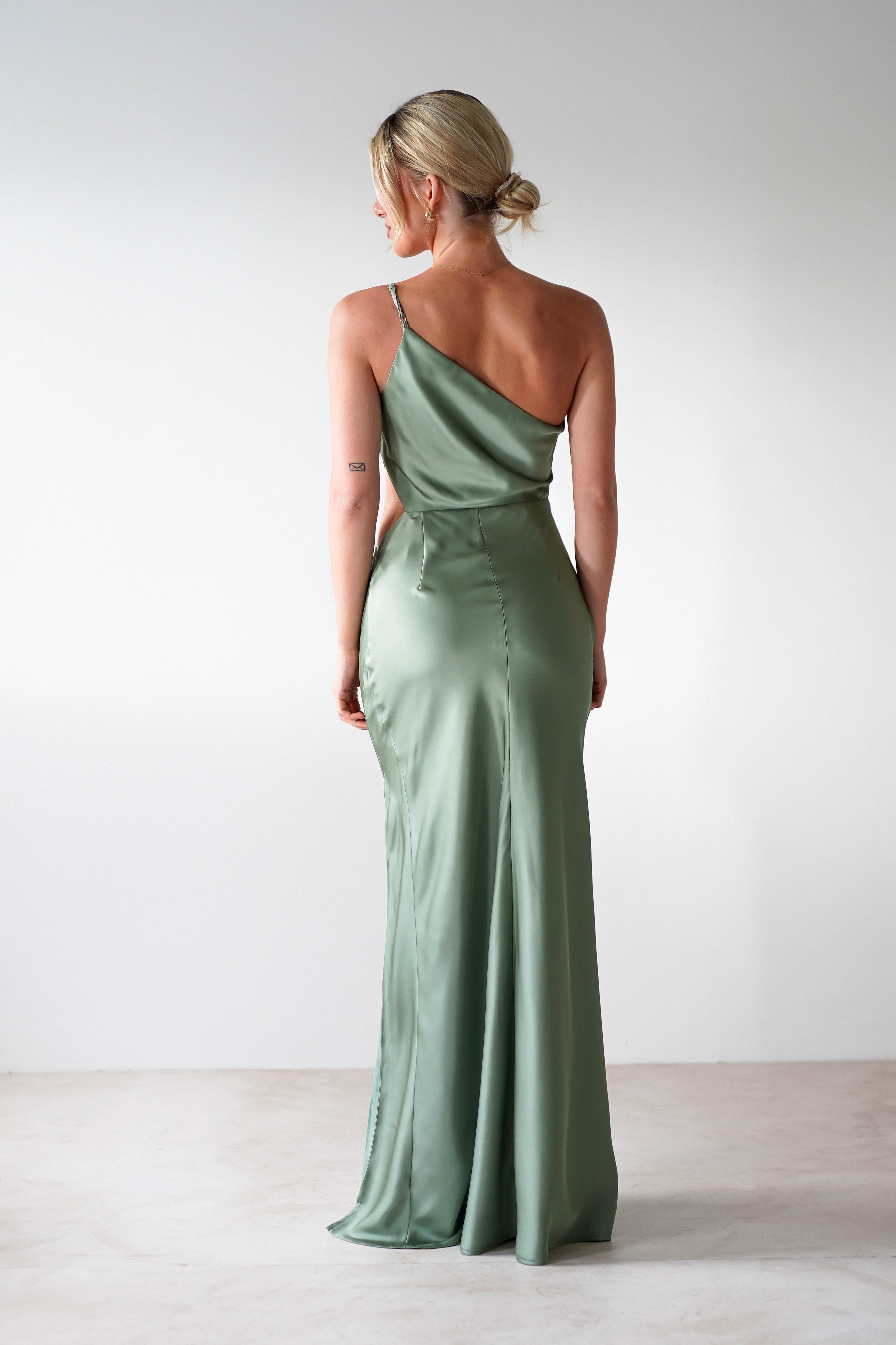 A person stands facing away, wearing a sleek, one-shoulder satin maxi in green with a floor-length hem. Their hair is styled in a neat bun. The background is plain and neutral, highlighting the elegant simplicity of the bridesmaid dress.