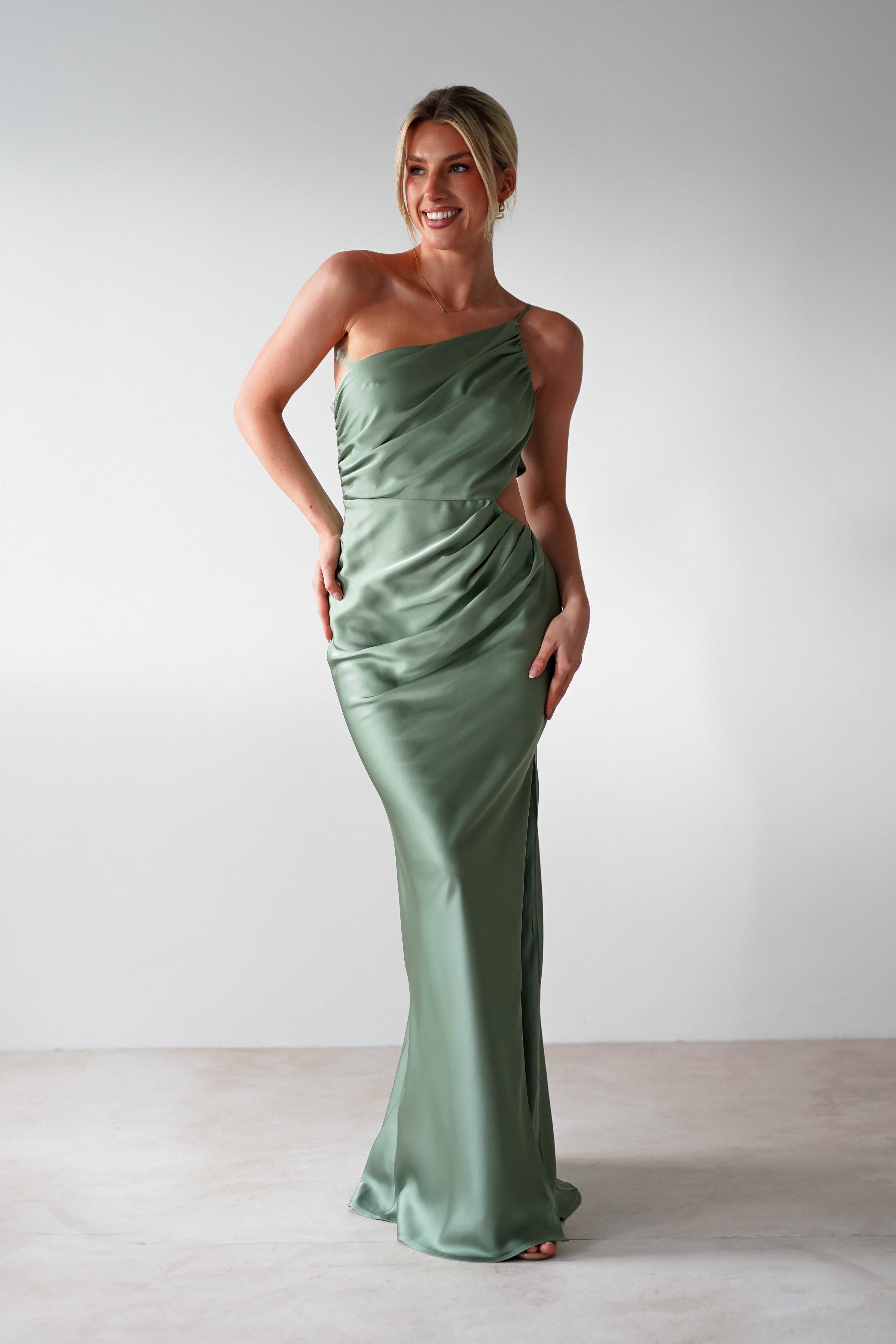 A person in a sleek, green satin maxi dress stands against a neutral background. The wedding guest dress is one-shouldered and draped elegantly. They are smiling and have their hair styled up.