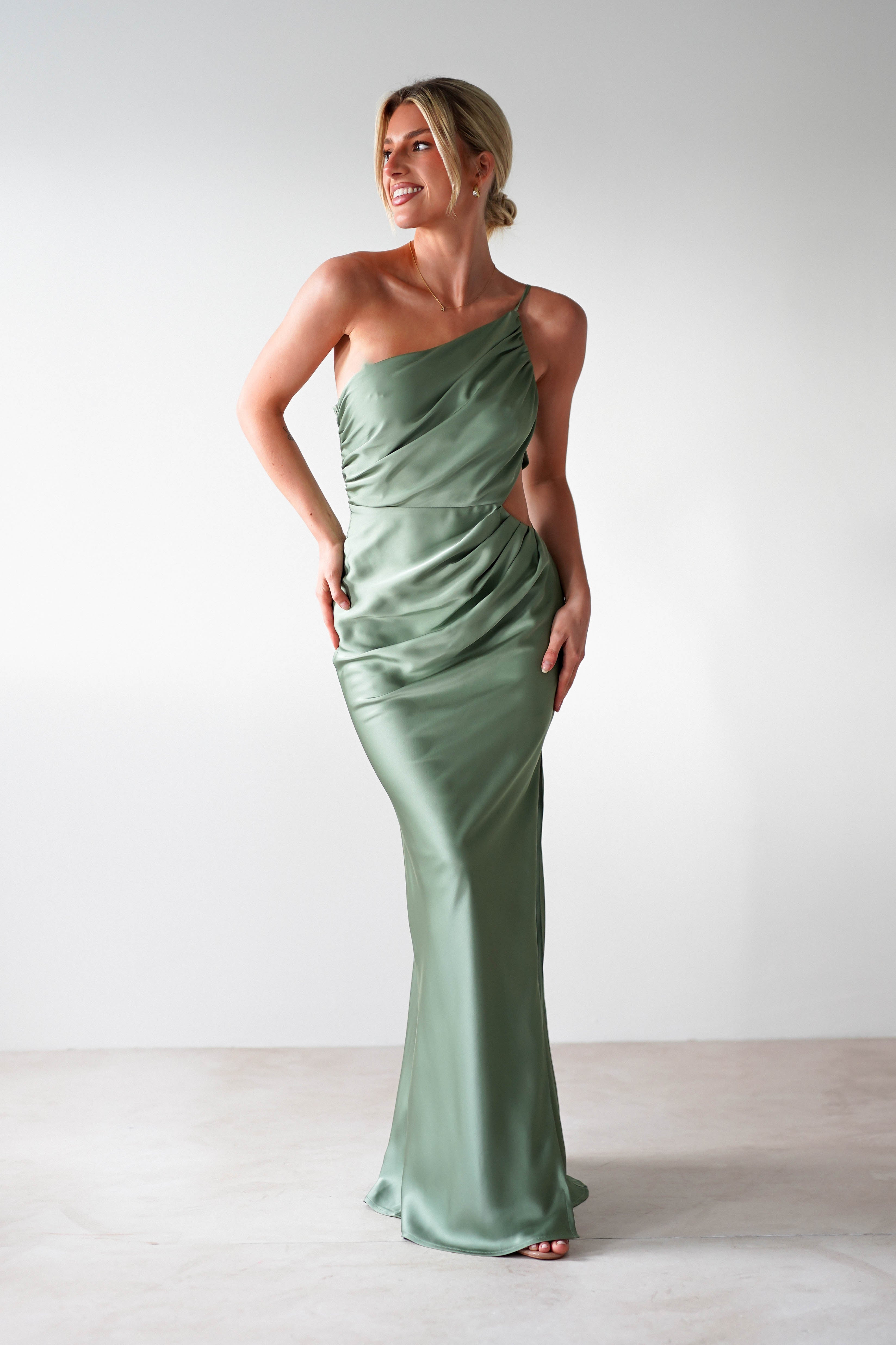 A woman in a green, one-shoulder satin maxi dress poses against a plain white backdrop. Her hair is tied back as she gazes to the side with a smile, exuding elegance perfect for a wedding guest.