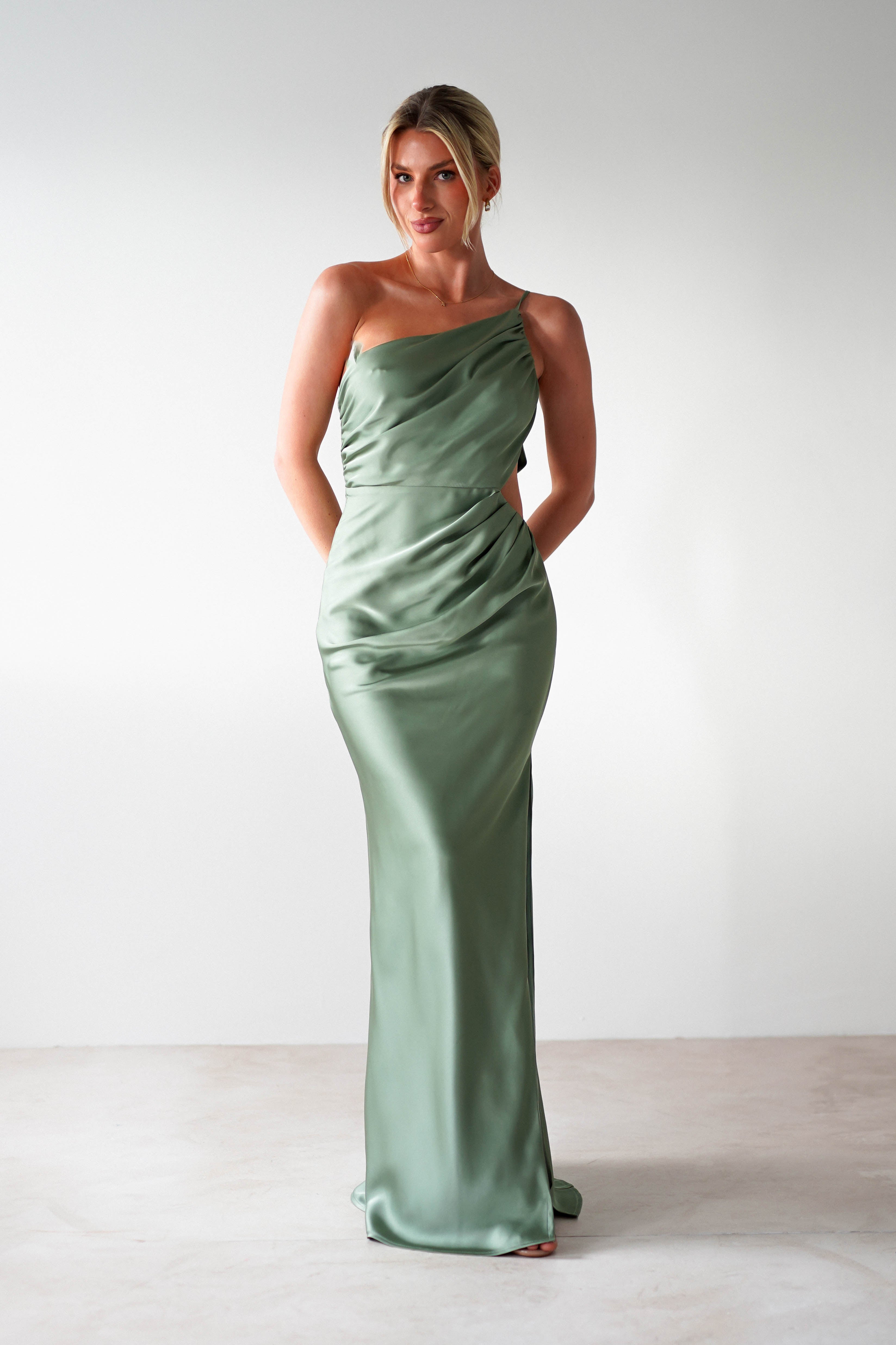 A woman stands in a minimalist setting wearing a mint green, one-shoulder satin maxi dress. Her hair is styled in an elegant updo, and she poses with her hands behind her back, embodying the perfect look for a wedding guest.