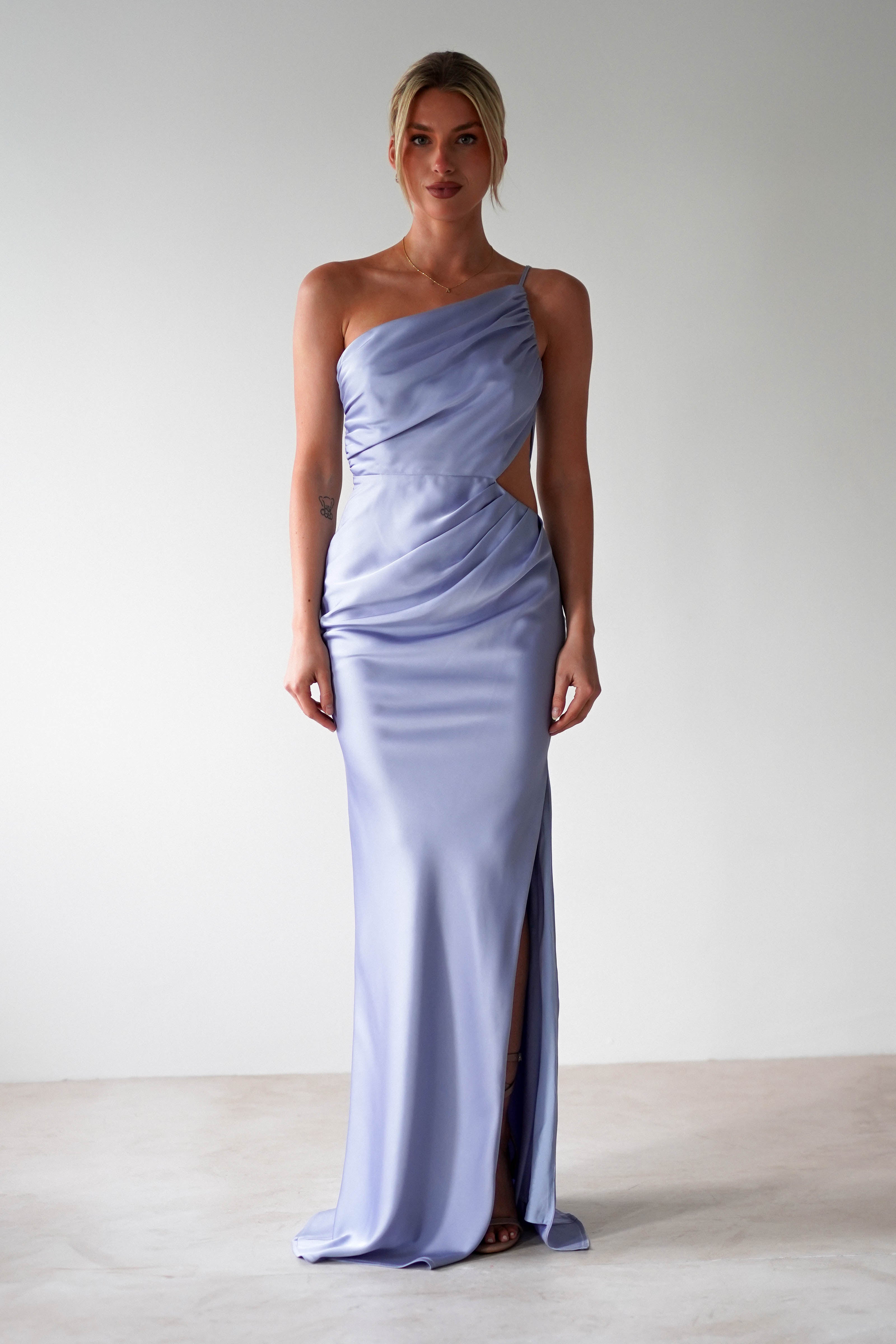 A woman stands against a plain light background wearing a silky one-shoulder satin maxi gown with draped detailing and a thigh-high slit—an elegant choice for bridesmaids or special occasions. Her hair is styled back as she faces forward.