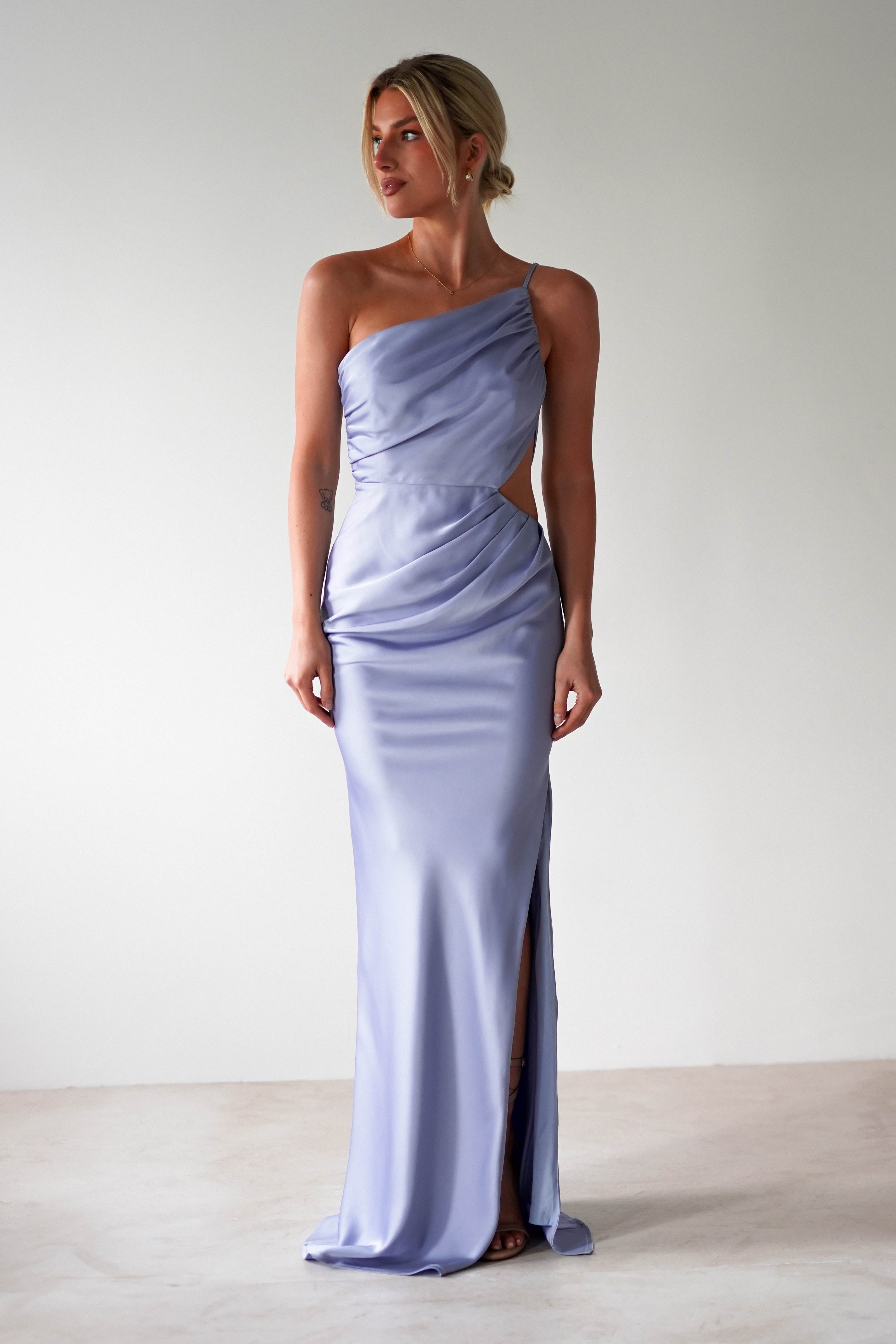 A woman wearing a matte satin blue one-shoulder satin maxi gown with a side slit stands against a plain, light-colored background. The dress features draped detailing and a fitted silhouette, perfect for bridesmaids.