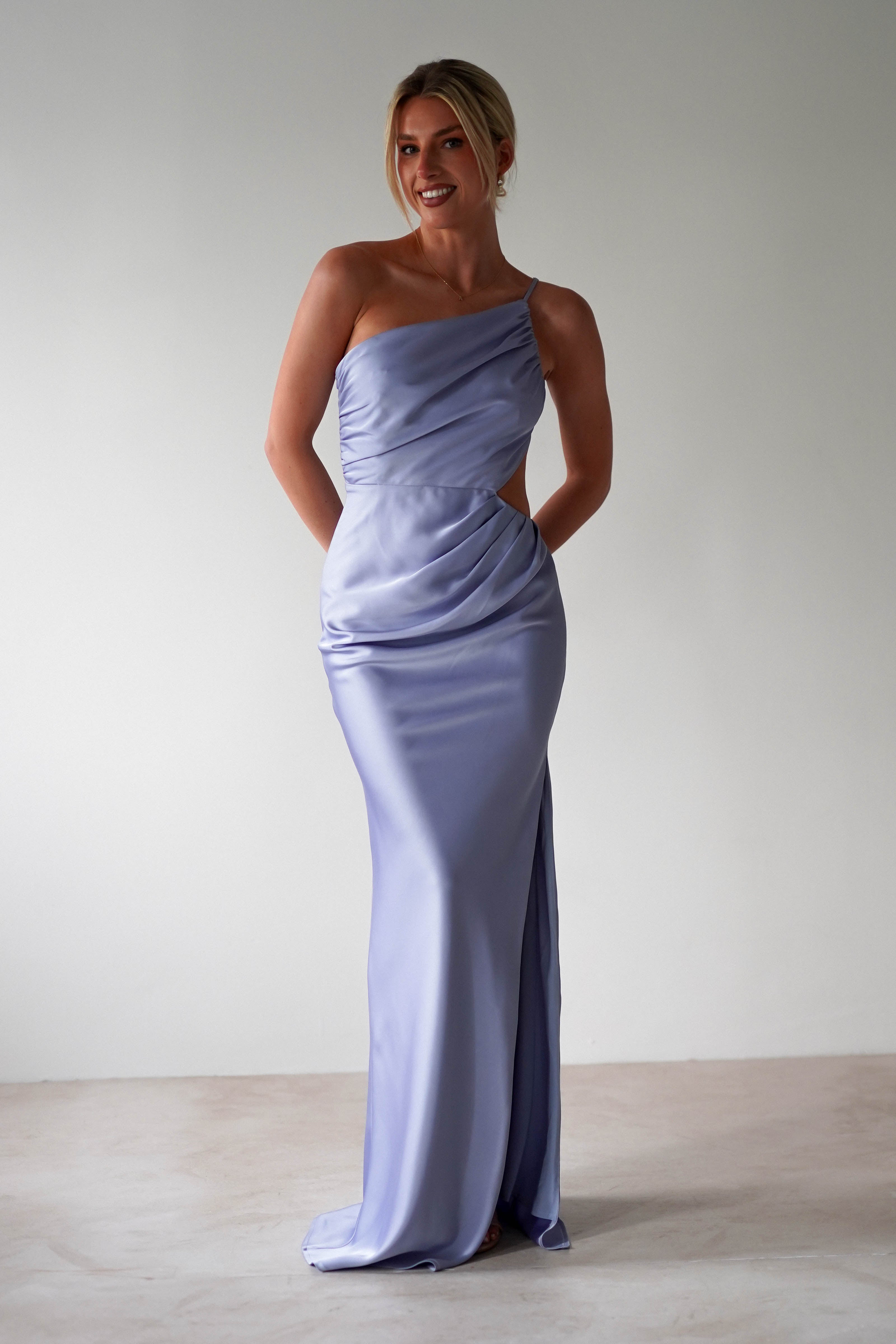A woman stands smiling, wearing a sleek, light lavender one-shoulder satin maxi gown with elegant draping and a fitted silhouette—perfect for bridesmaids—while she poses against a plain, light-colored background.