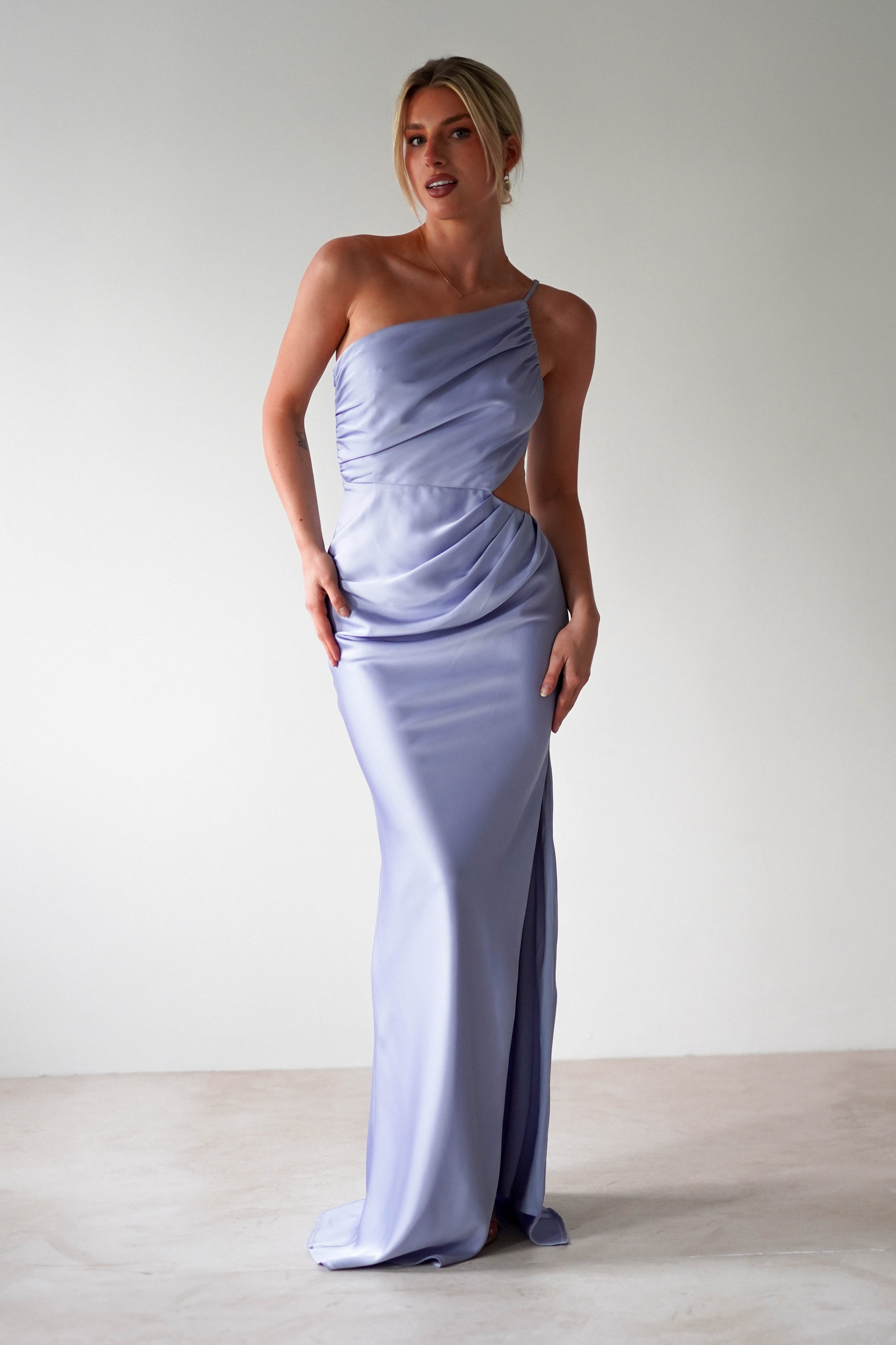A woman wearing a matte satin blue, one-shoulder satin maxi gown stands against a plain white background, posing with her hands resting on her hips and side—an elegant look perfect for bridesmaids.